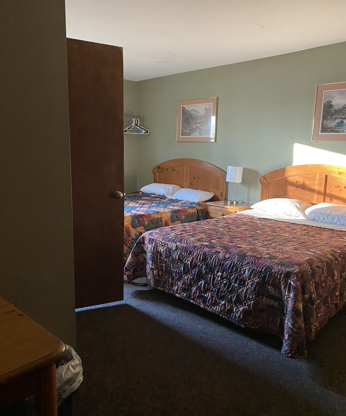 A hotel room with two beds, wooden headboards, a nightstand with a lamp, and framed landscape pictures on the green walls. One bed has a multicolored quilt, and the other has a darker quilt with a pattern. An open door partially obscures the view.