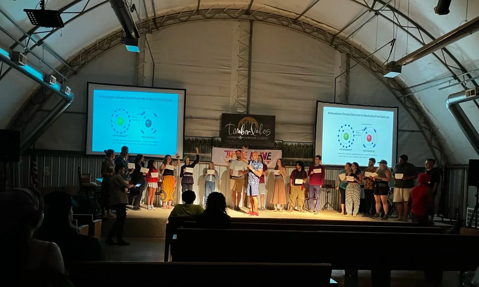 People standing on a stage during a presentation or event inside a large tent, with two large projection screens displaying diagrams about antioxidants donating electrons to neutralize free radicals.