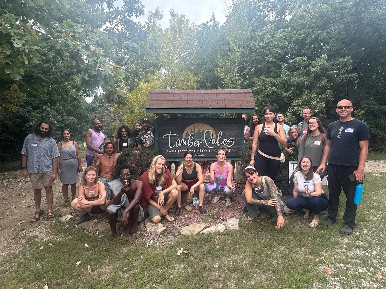 Group of people gathered outdoors around a sign that reads 'Timber Lakes camp retreat center'. They are smiling and posing for a photo, with some sitting and others standing, in a wooded area.