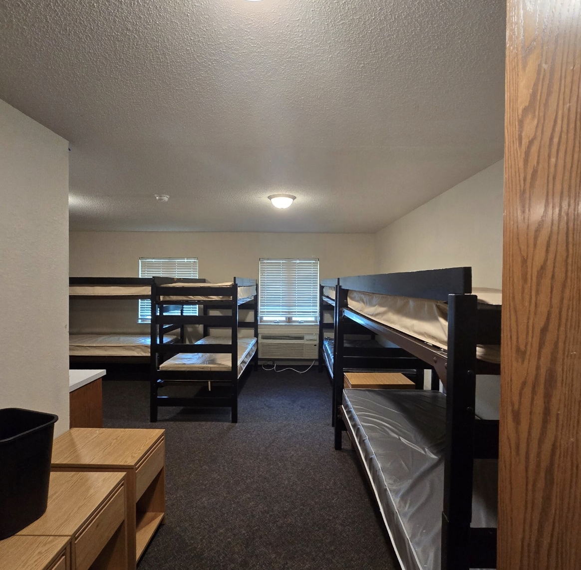 Dormitory room with two sets of black bunk beds, three windows with blinds, an air conditioning unit, a wooden furniture piece, and a waste basket.