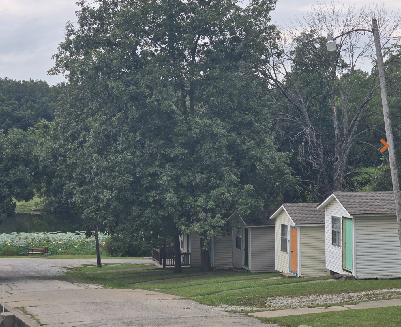 Small houses with colorful doors in a park-like setting near a body of water, with trees and a bench in the background, overcast sky.