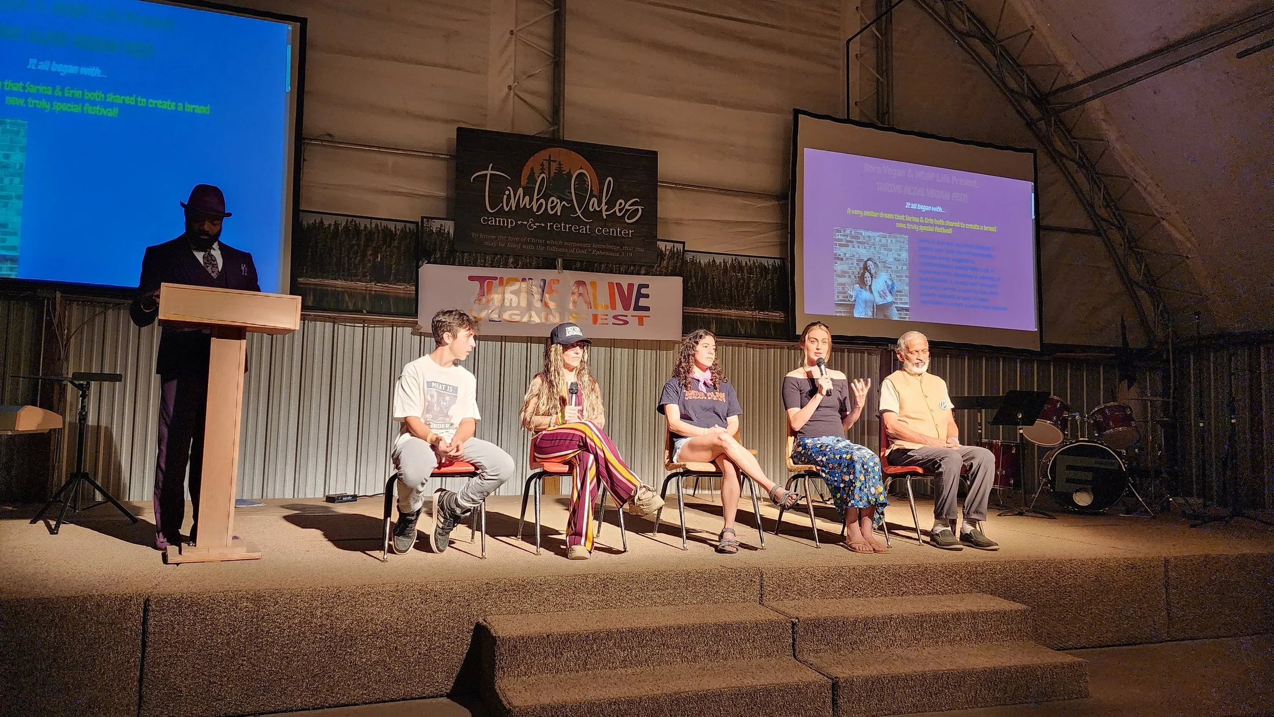 Panel discussion on stage at Timberlakes Camp & Retreat Center during TRIPLE ALIVE FEST. Six people are seated in a row, five women and one man, with a man standing at a podium on the left. Two large screens display a presentation behind them.