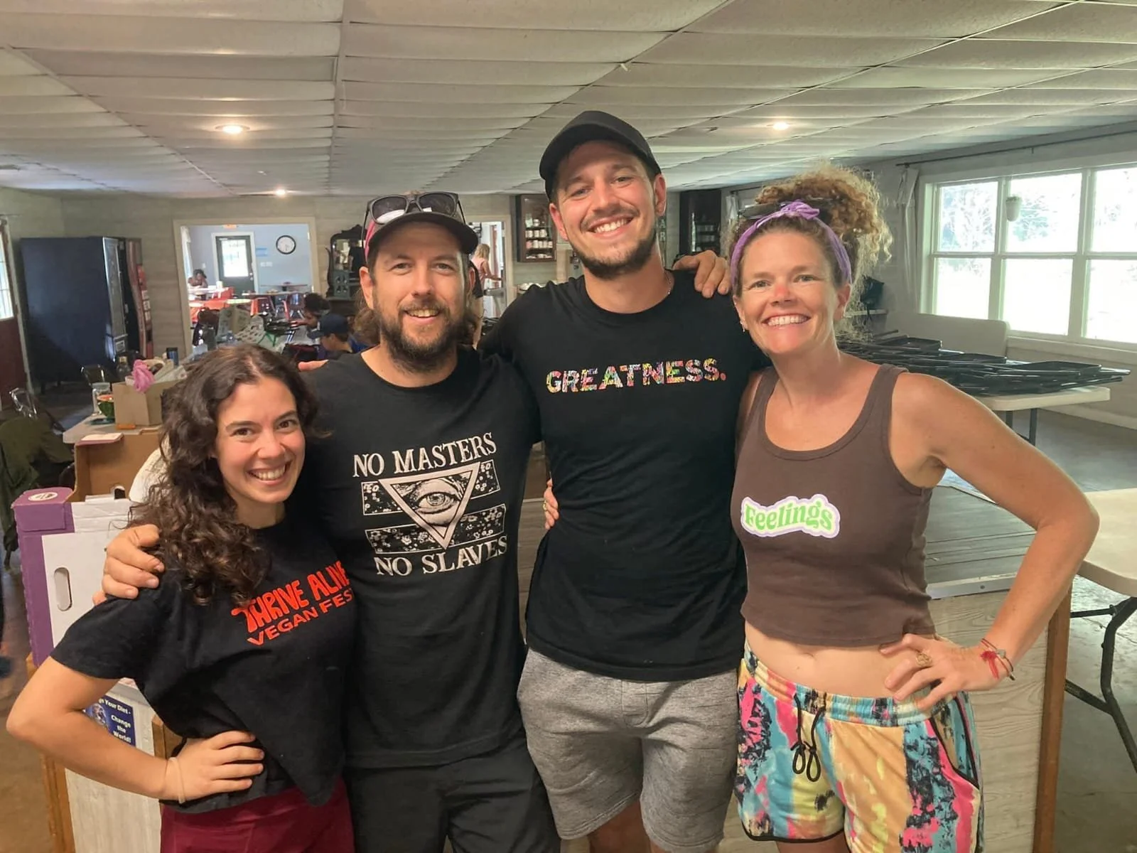 Four smiling people standing indoors, embracing each other. The group includes two women and two men, all casually dressed. The background shows a room with tables, chairs, and large windows.