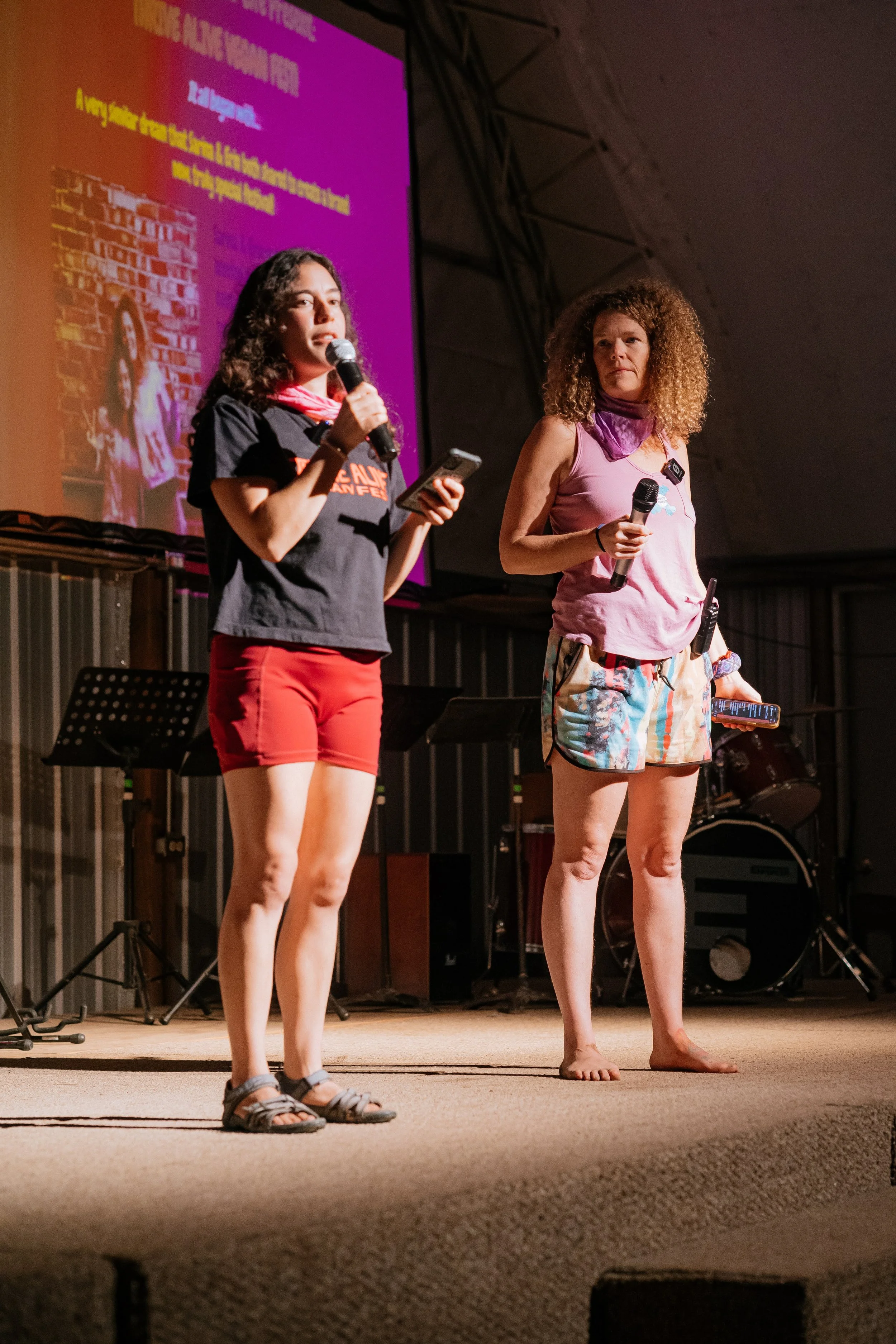 Two women standing on stage holding microphones, one speaking and the other listening, with a large pink screen behind them displaying text.