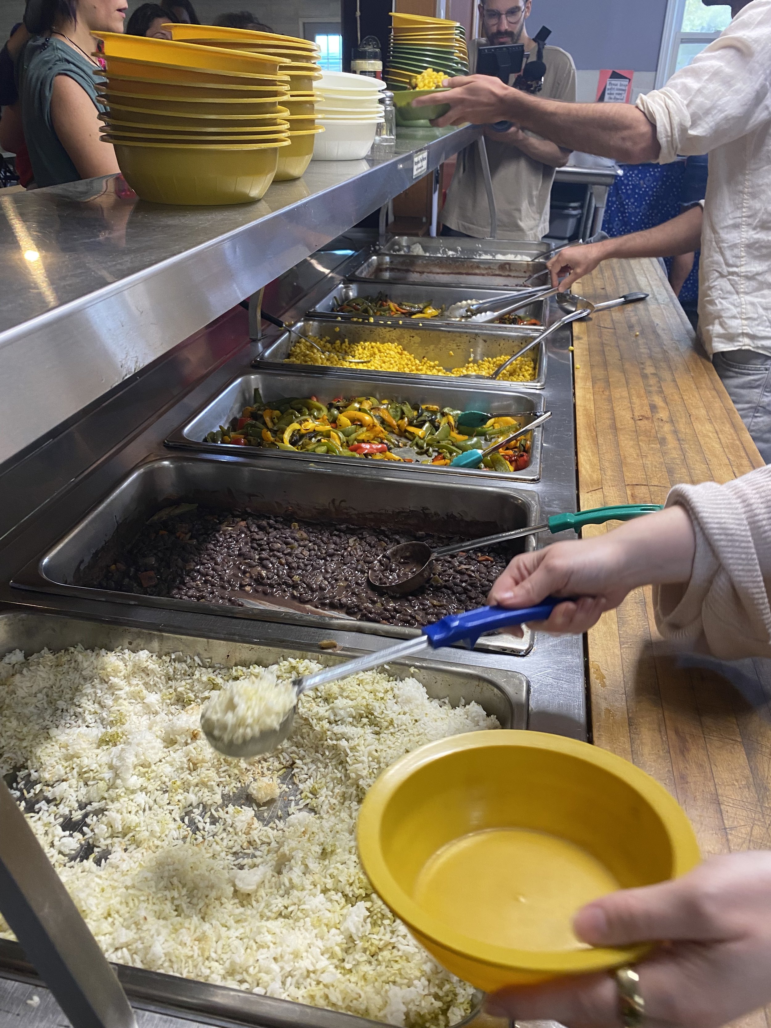 Buffet with various dishes including black beans, rice, yellow corn, mixed vegetables, and other hot foods, with people serving themselves.