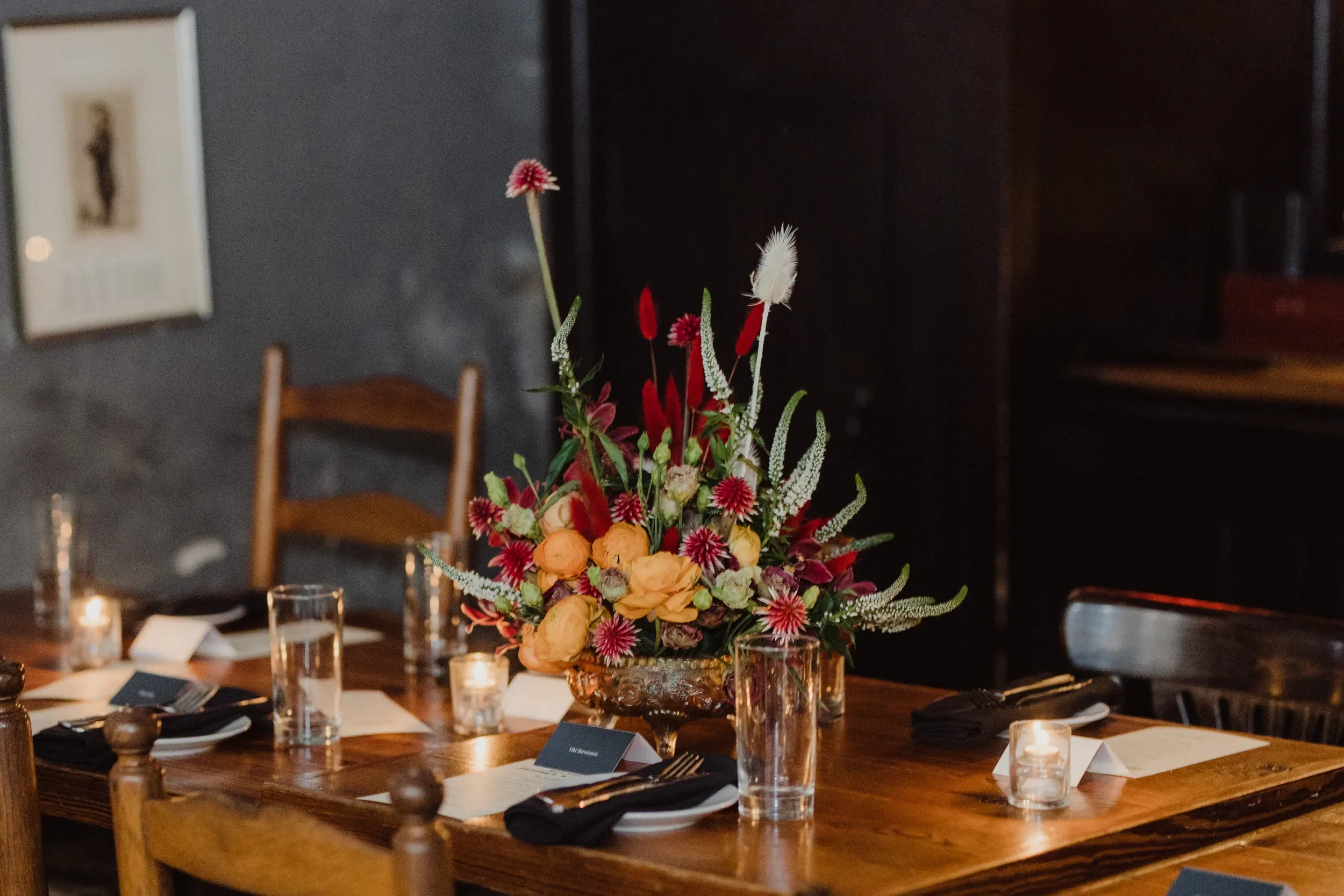 A dining table decorated with a large colorful floral centerpiece, surrounded by glasses, napkins, and lit candles in a dimly lit room.