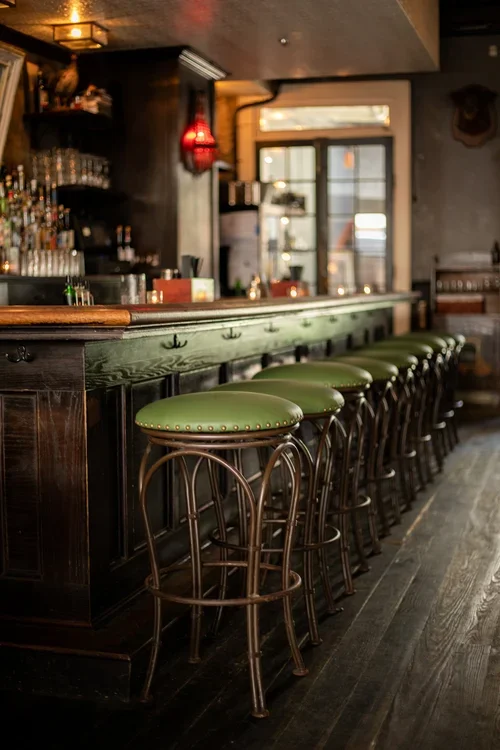 Empty bar with green cushioned stools at a wooden counter in a dimly lit pub.