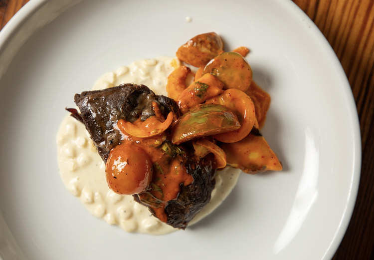 A plate of cooked meat with tomato-based sauce, served over a bed of white creamy substance, garnished with sliced vegetables.