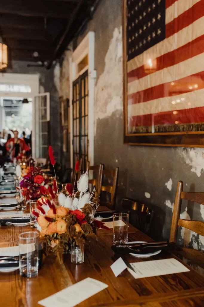 A long wooden dining table set for a meal with black plates, clear glasses, and place cards. The table has floral centerpieces with red, orange, and white flowers. The restaurant has a rustic look with a partially worn wall and an American flag frame