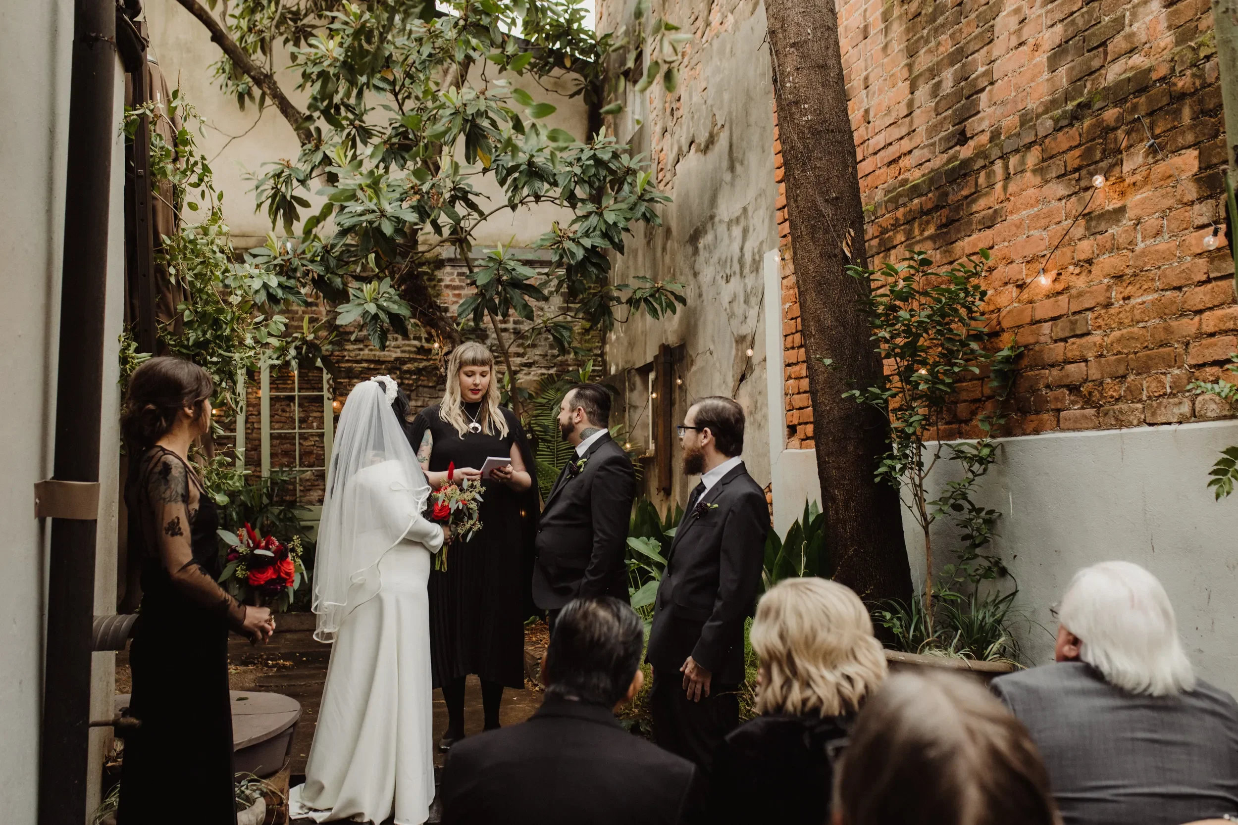 A wedding ceremony taking place in an intimate outdoor courtyard with brick and concrete walls, trees, and string lights. The bride and groom stand before an officiant, with bridesmaids and groomsmen nearby, and guests seated watching.