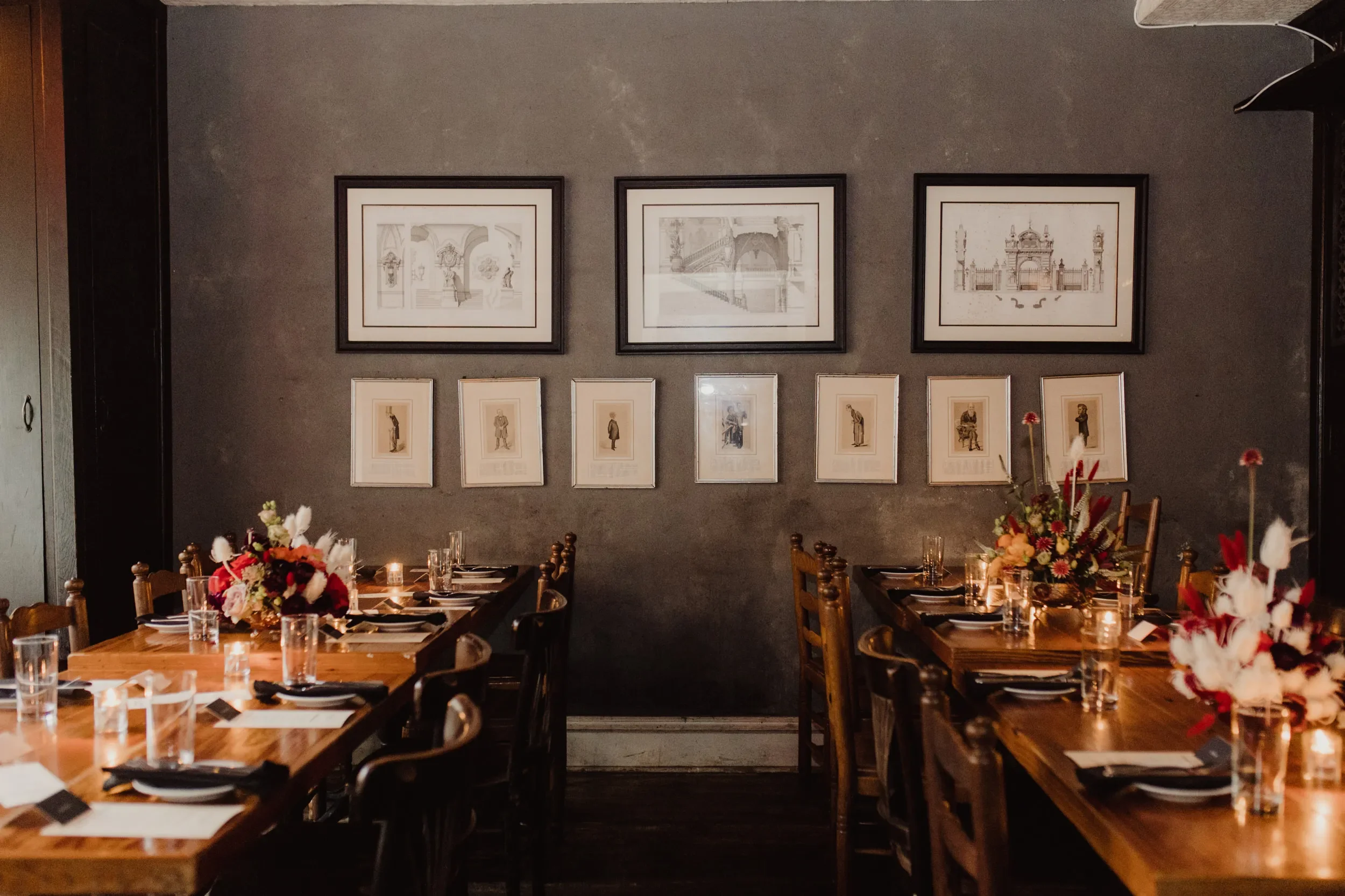A dining room with two long wooden tables set with black plates, clear glasses, and candlelit centerpieces with colorful flowers. The room has a dark gray wall decorated with framed artwork.