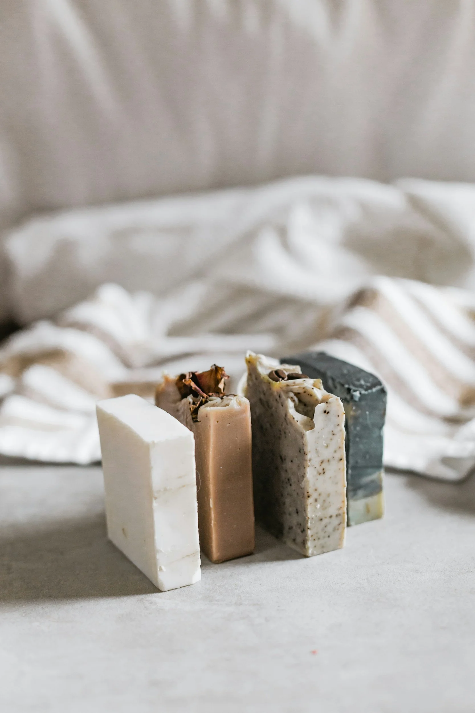 Four bars of handmade soap in different colors and textures, arranged in a row on a neutral surface with a light-colored, textured fabric in the background.