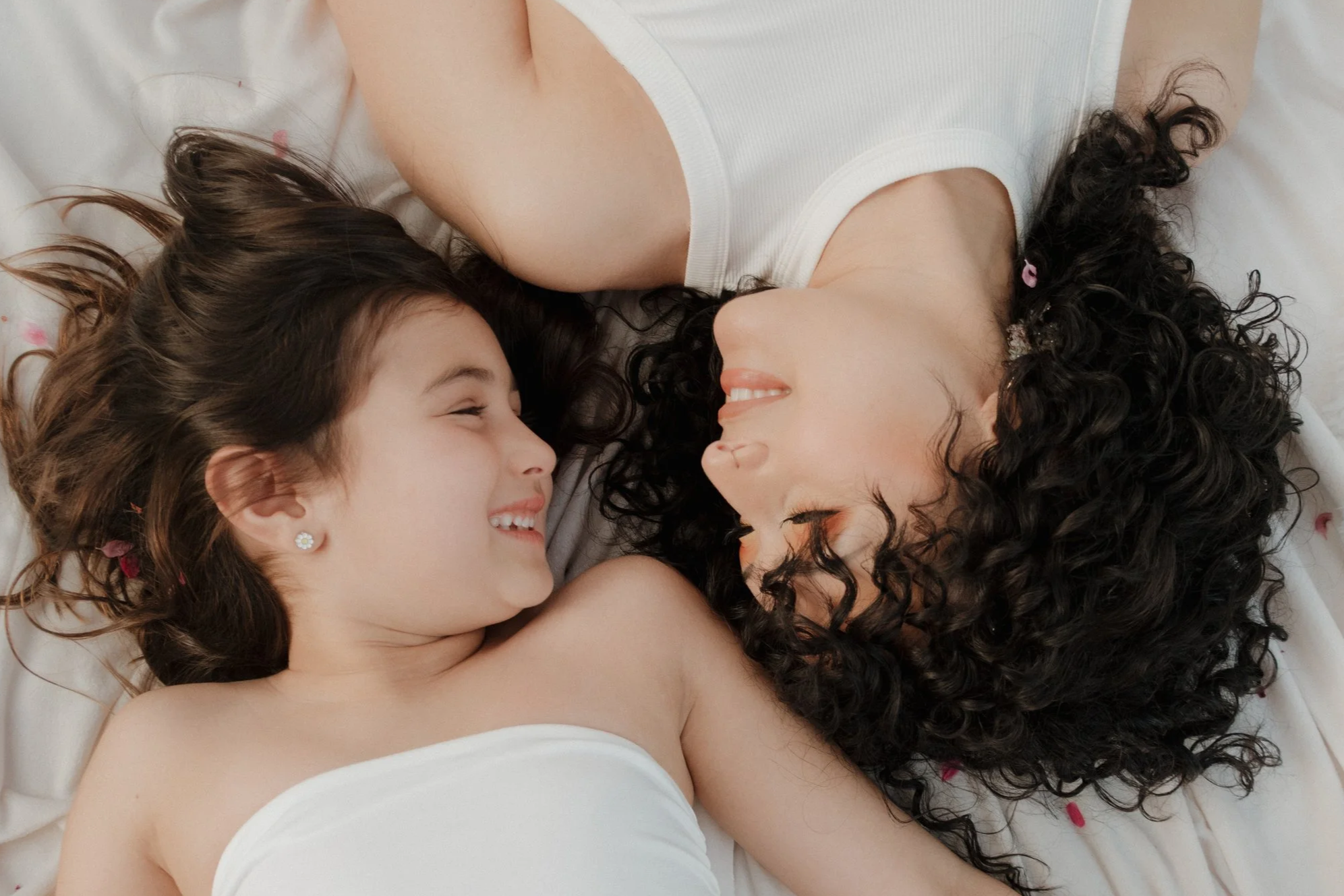 A smiling woman and a girl lying on a bed, facing each other with their heads close together, both wearing white sleeveless tops and smiling.