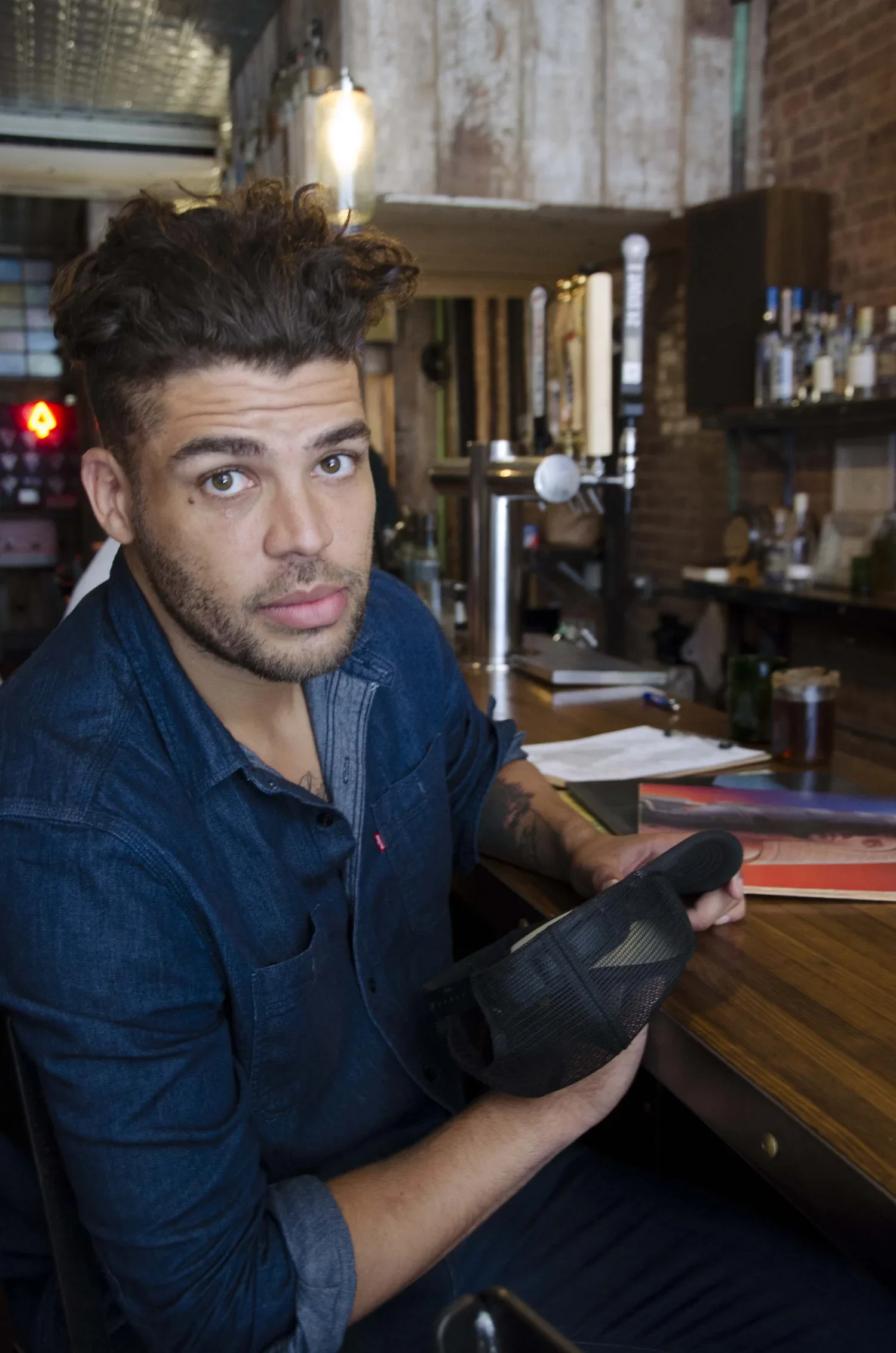 A young man with brown, curly hair and a beard sitting at a bar counter inside a rustic restaurant or bar with exposed brick walls. He is holding a small black mesh bag and looking at the camera.