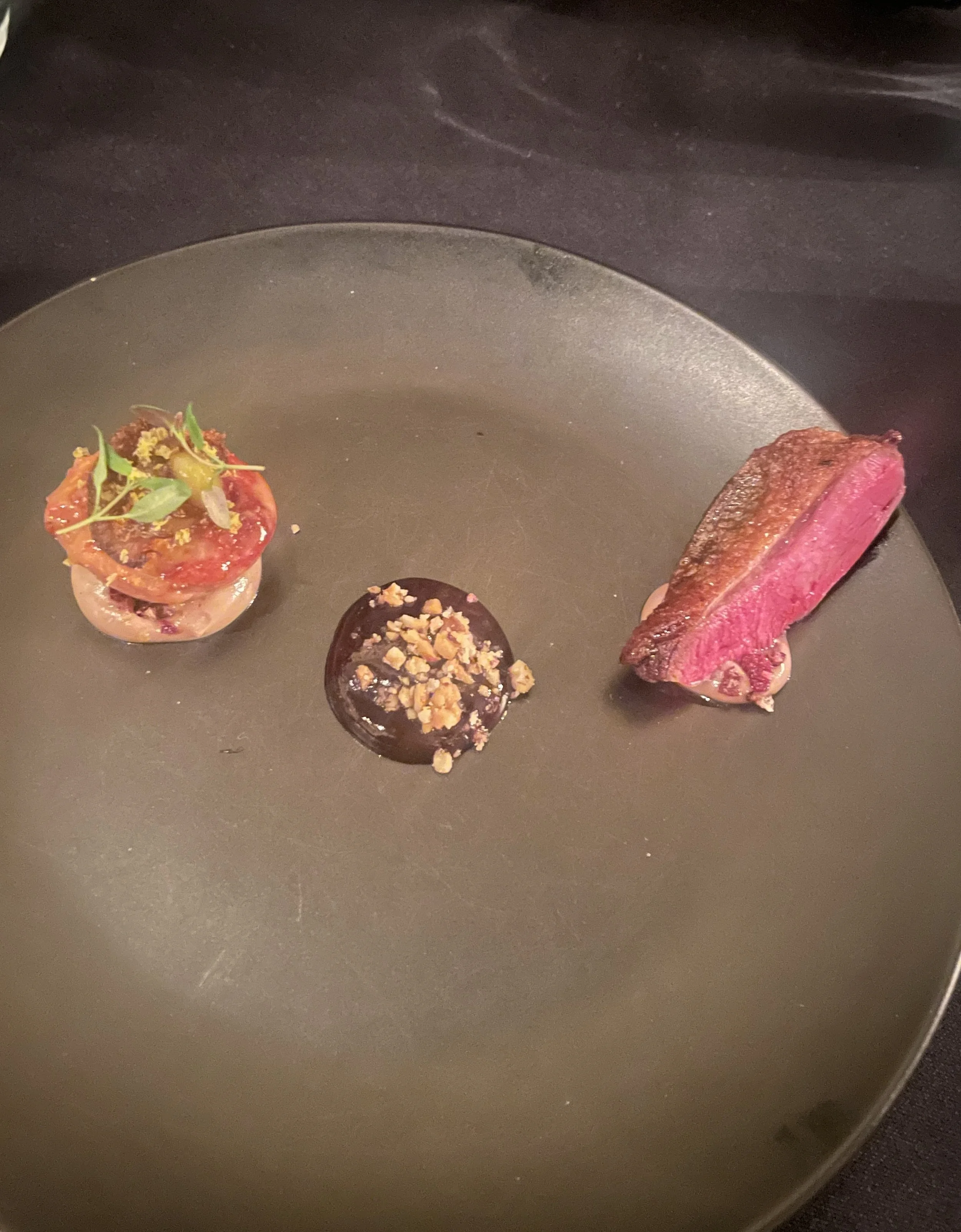 A gourmet dish with three small, artfully arranged food items on a dark, round plate. The items include a piece of medium-rare steak, a small round chocolate dessert with crushed nuts on top, and a bite-sized portion topped with microgreens and a che