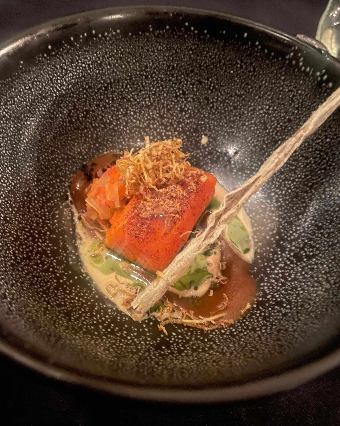 A black textured bowl containing a piece of cooked carrot topped with herbs, a sprig of sage, and some sautéed vegetables in broth.