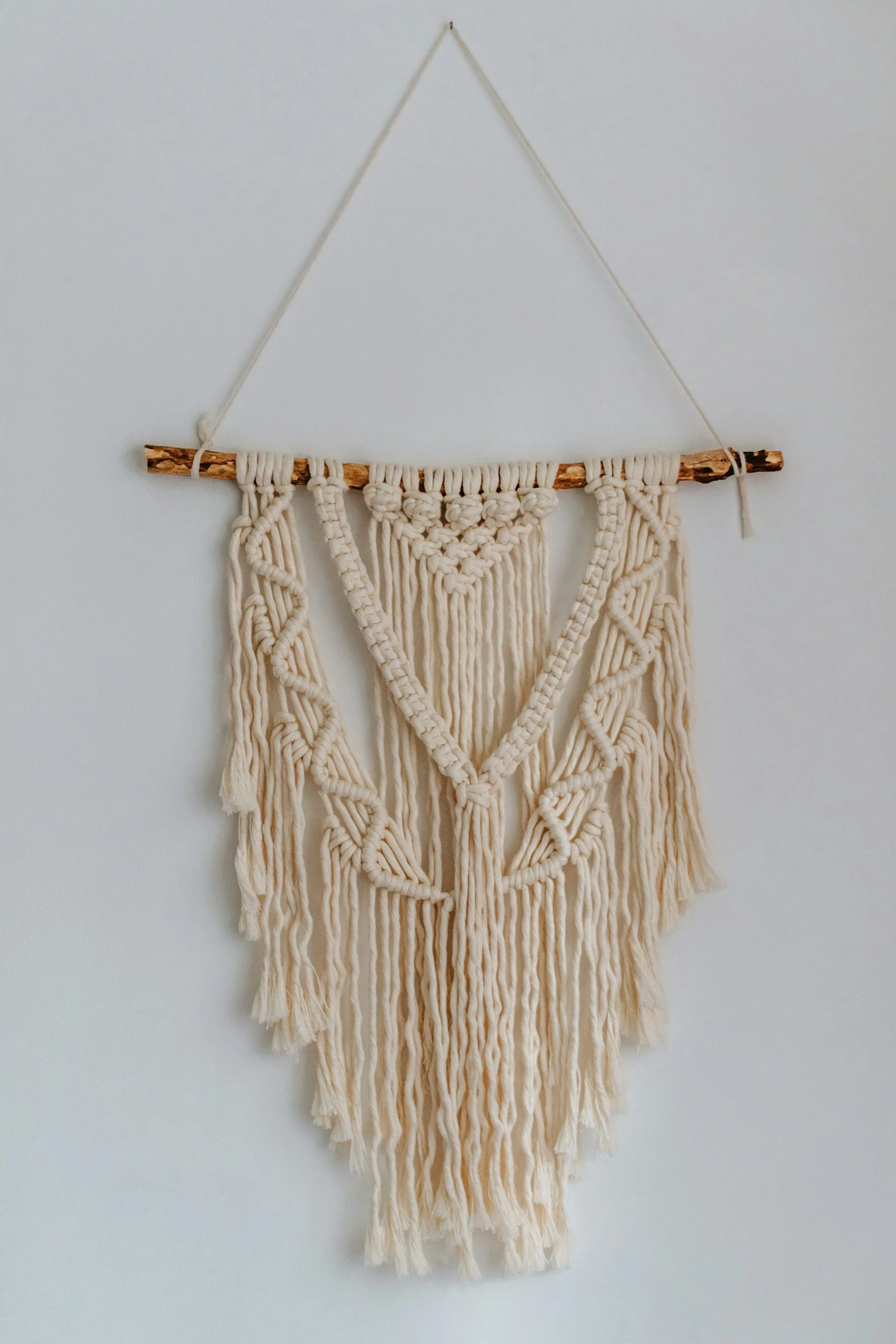 Beige macrame wall hanging with a wooden rod and hanging string, featuring intricate woven patterns and fringe at the bottom, displayed against a plain white wall.