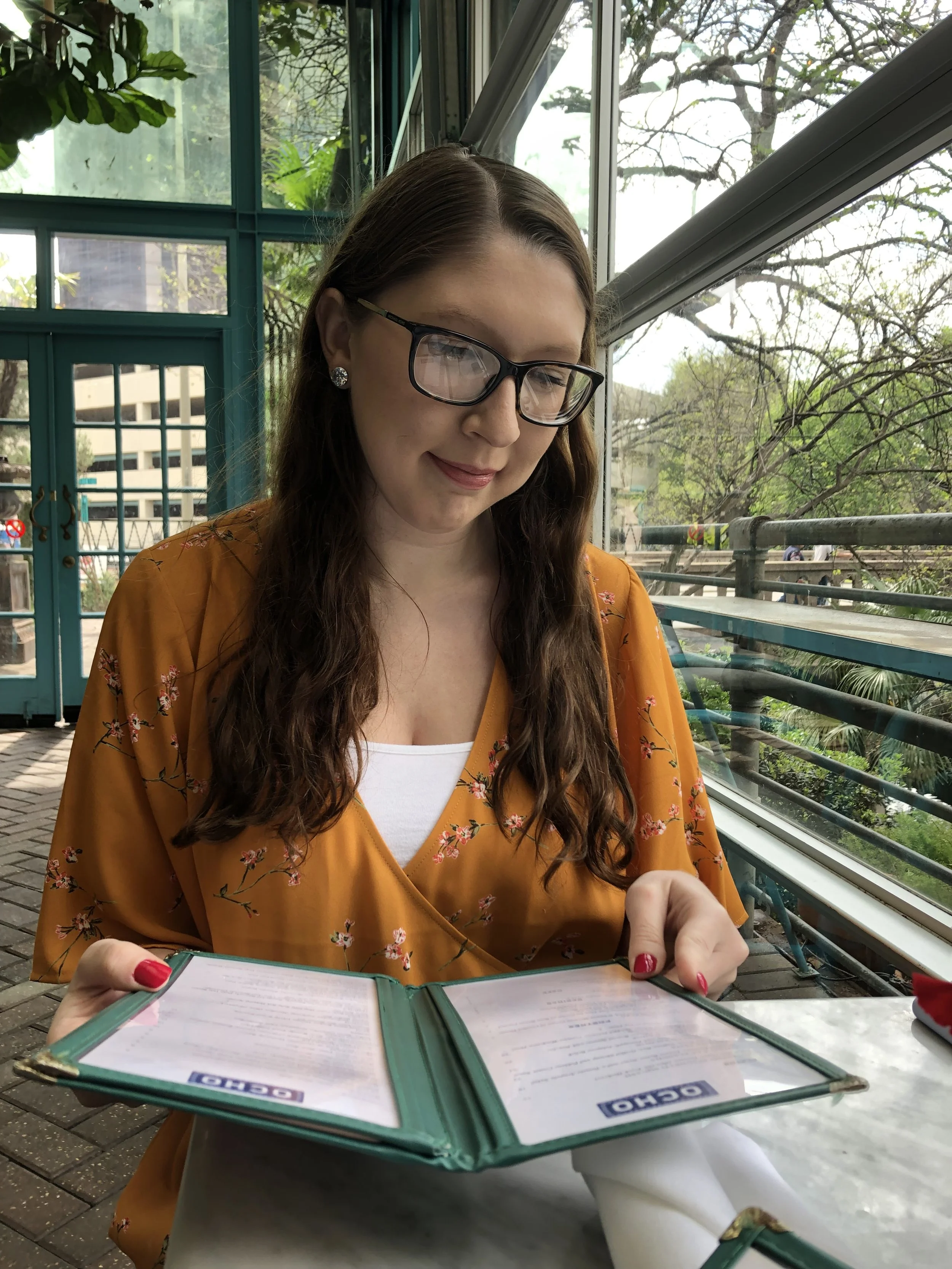 A young woman with long brown hair, glasses, and earrings, wearing a mustard yellow floral blouse, is sitting at a table inside a glass-walled restaurant or cafe, looking at a menu.