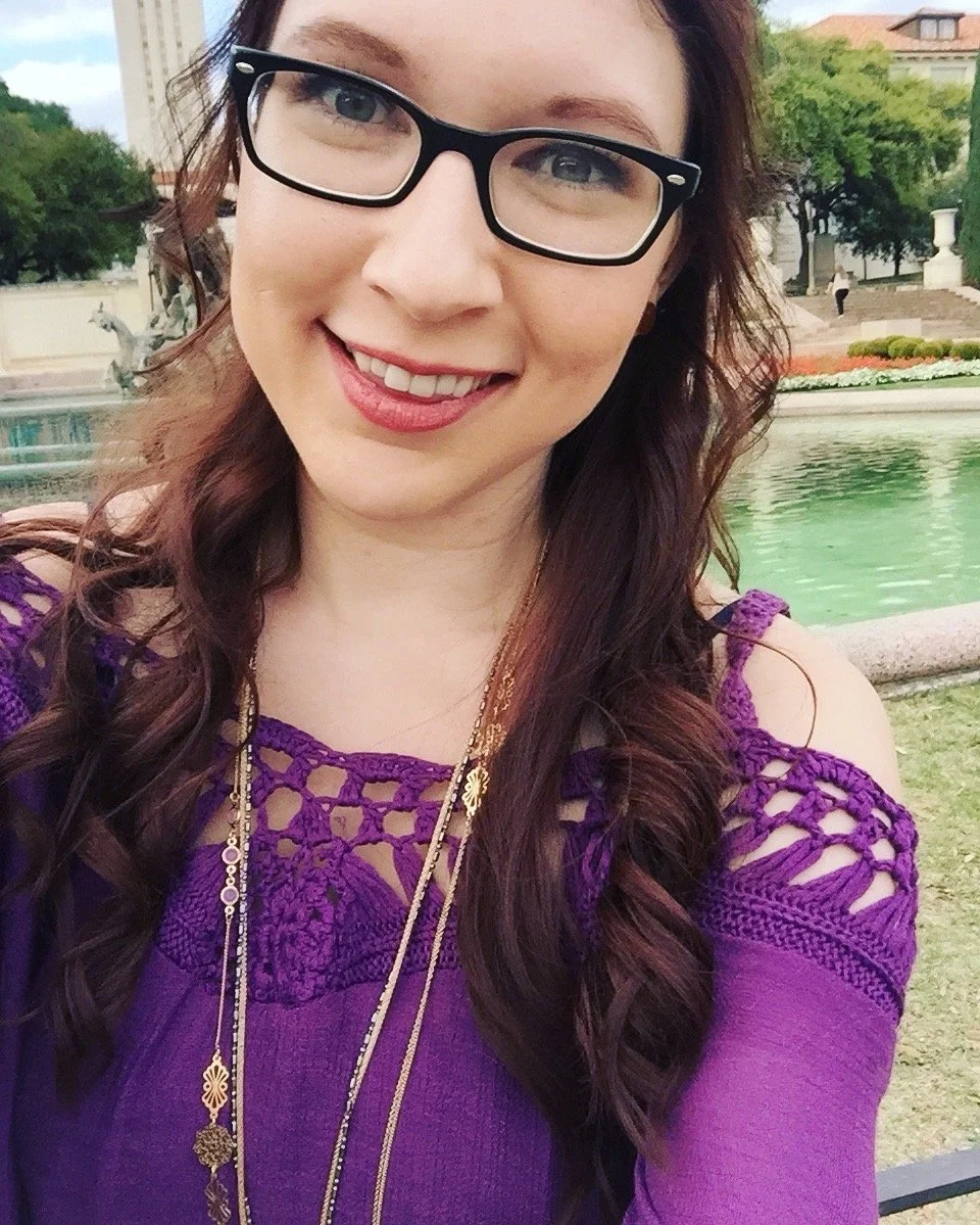 A young woman with long, wavy brown hair wearing glasses and a purple crochet top, smiling outdoors near a fountain and greenery.