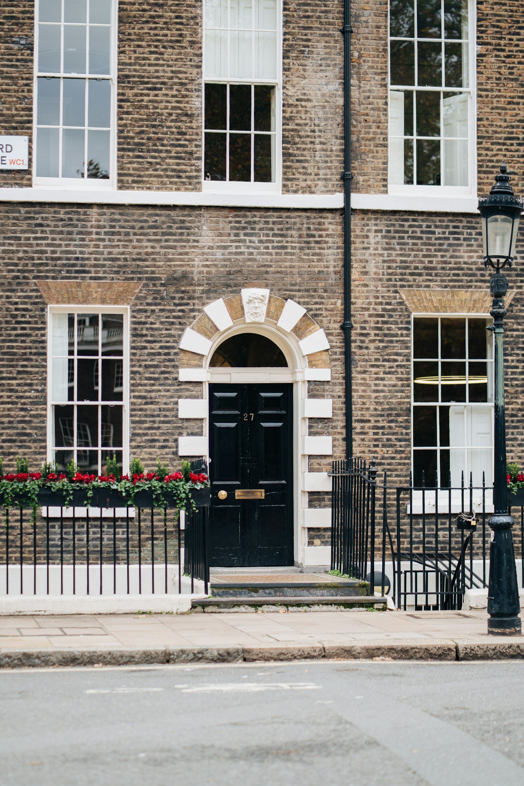 A brick building with a black front door numbered 27, surrounded by white stone accents, three white-framed windows, a flower box with red flowers, and a black street lamp to the right.