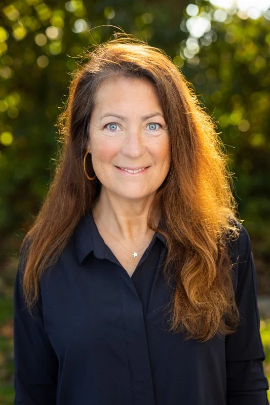 A woman with long brown hair, wearing a navy blue shirt and gold hoop earrings, smiling outdoors with a blurred green background.