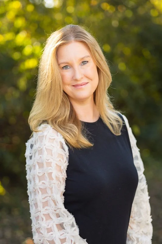 A woman with long blonde hair smiling outdoors in natural light, wearing a black top with white lace sleeves.