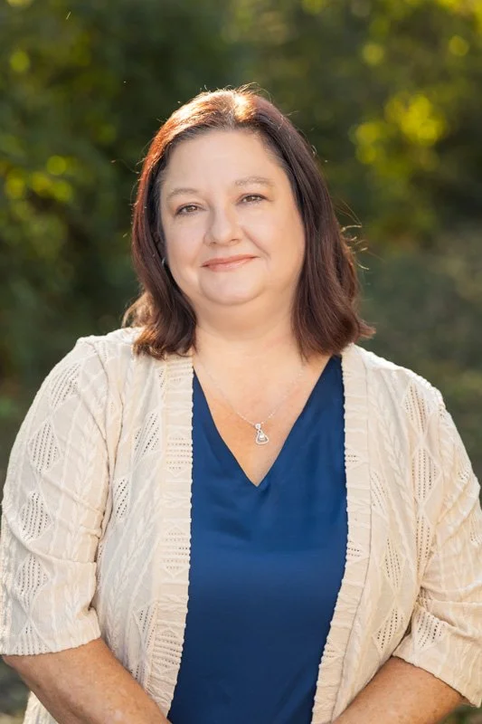 A woman with shoulder-length brown hair and fair skin, wearing a navy blue top and a light-colored, open-knit cardigan, smiling outdoors with blurred green foliage in the background.