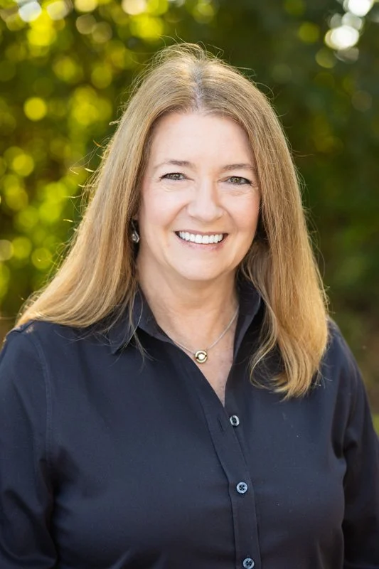 A smiling woman with long blonde hair wearing a navy blue button-up shirt standing outdoors with blurred green foliage in the background.