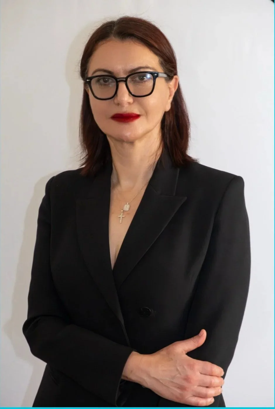 A woman with shoulder-length dark hair, wearing glasses, a black blazer, and red lipstick, standing against a white wall.