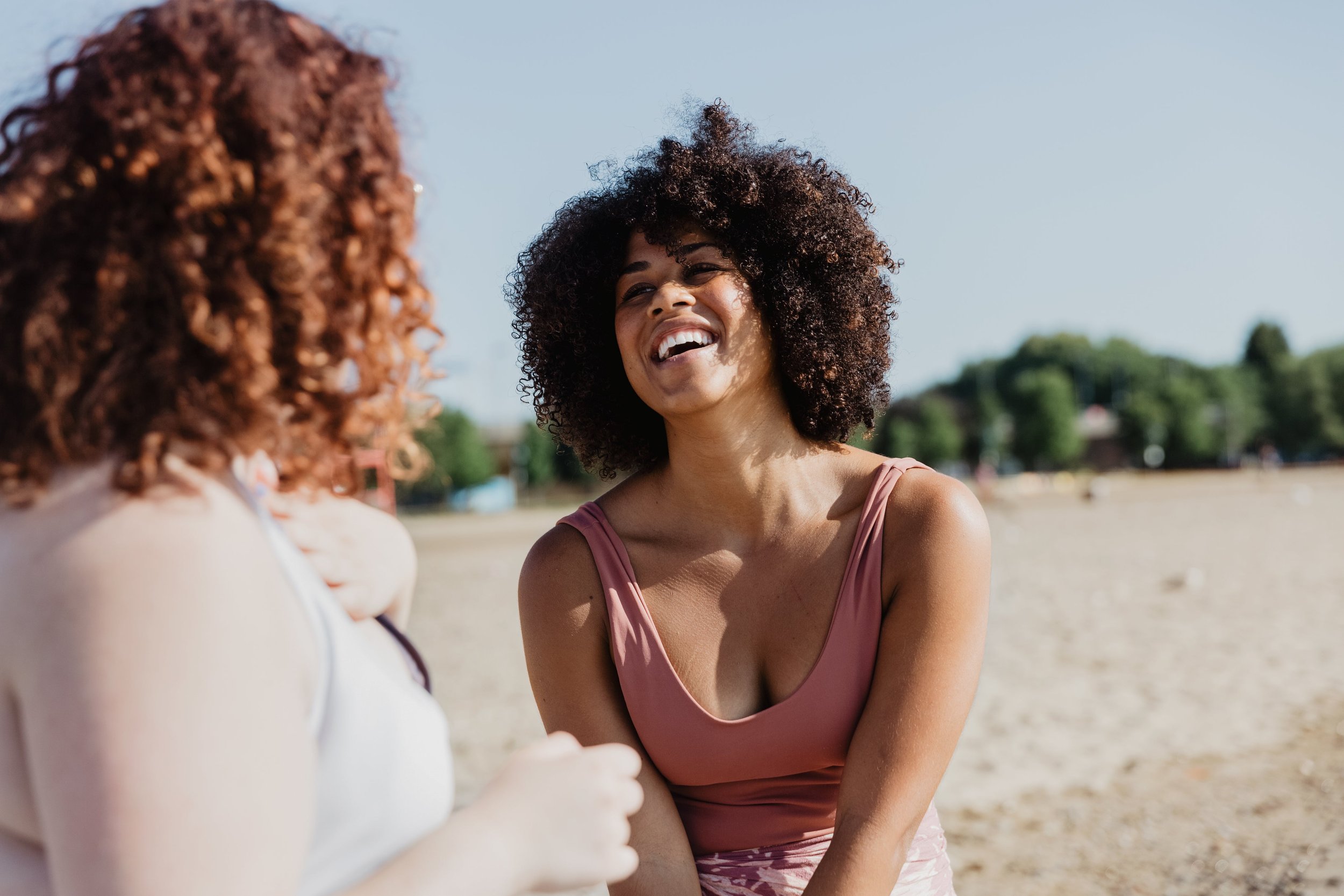 Two women with curly hair smiling and engaging in conversation outdoors on a sunny day.