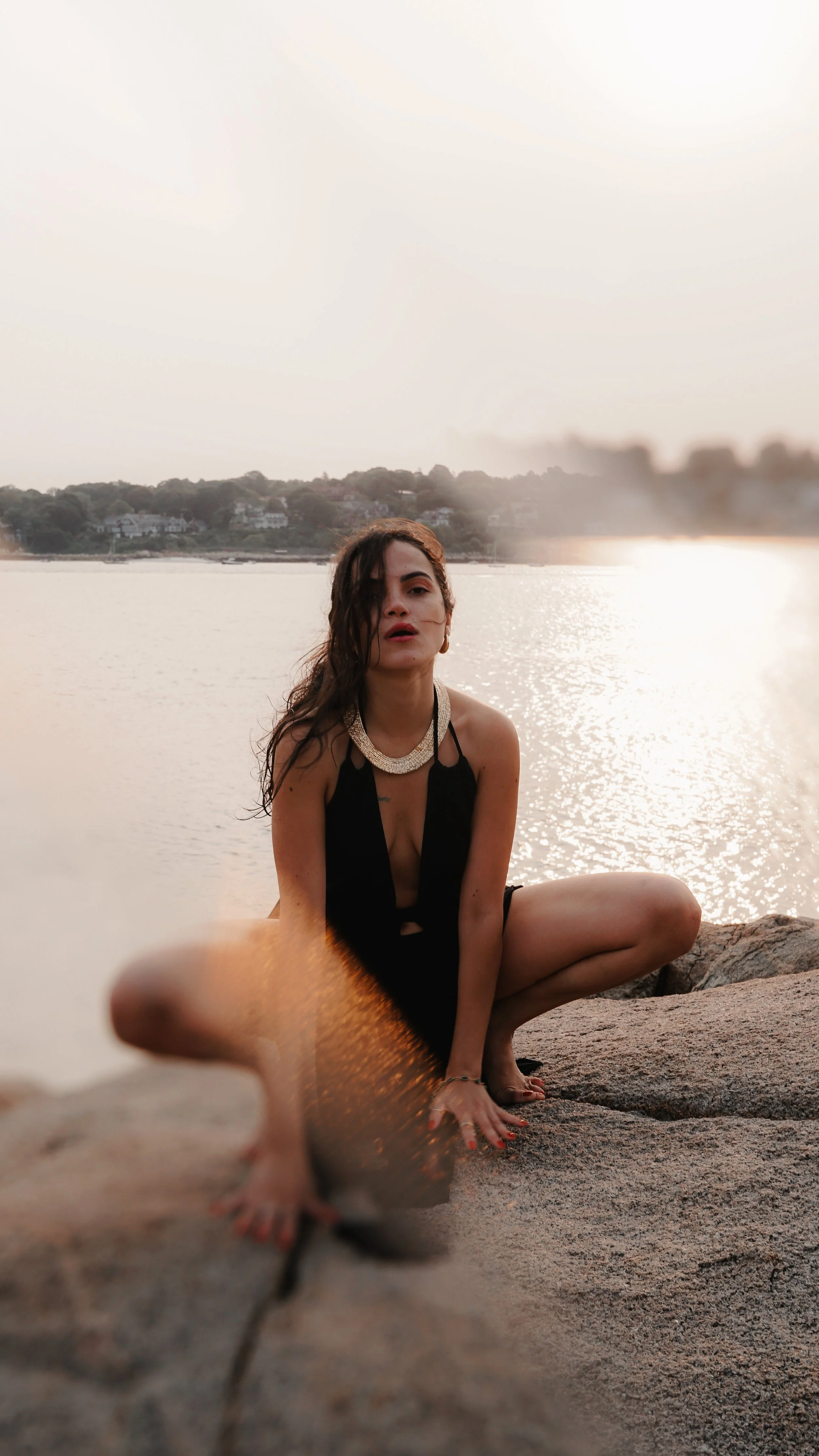 A woman in a black dress with a necklace is sitting on rocks by a body of water during sunset, with her hair partly covering her face.