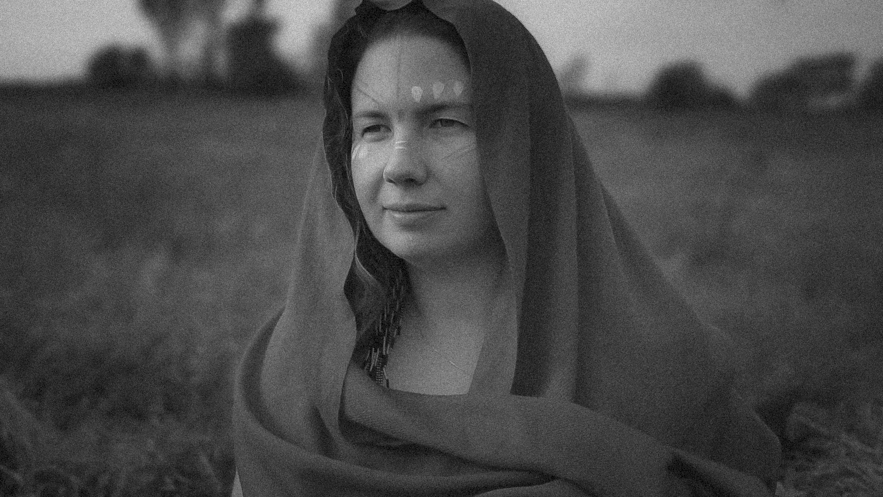 Black and white photo of a woman with long hair, wrapped in a shawl or cloak, outdoors in an open field with blurred trees in the background.