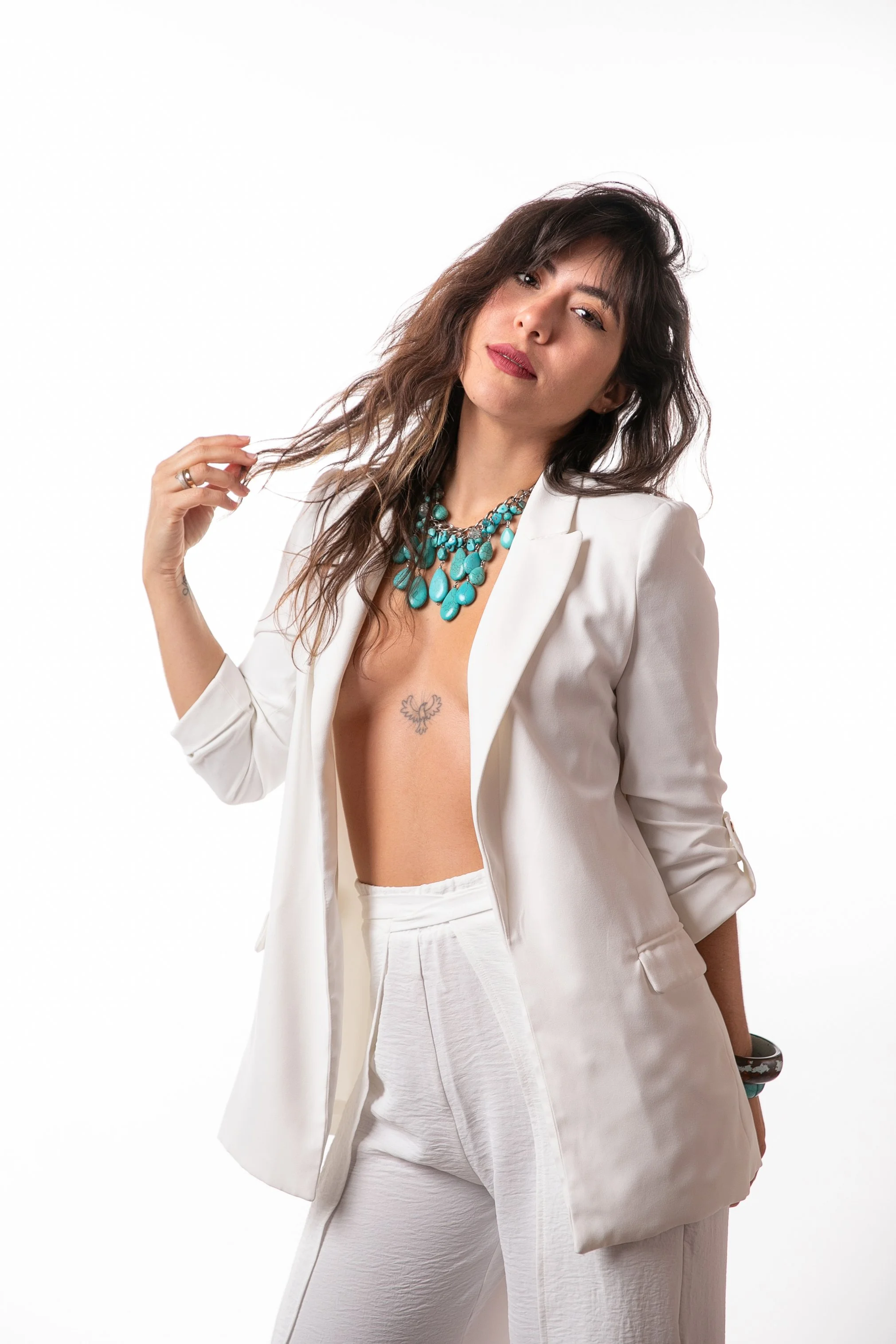 A woman with wavy brown hair, wearing a white blazer and white pants, accessorized with a turquoise necklace and rings, posing against a plain white background.