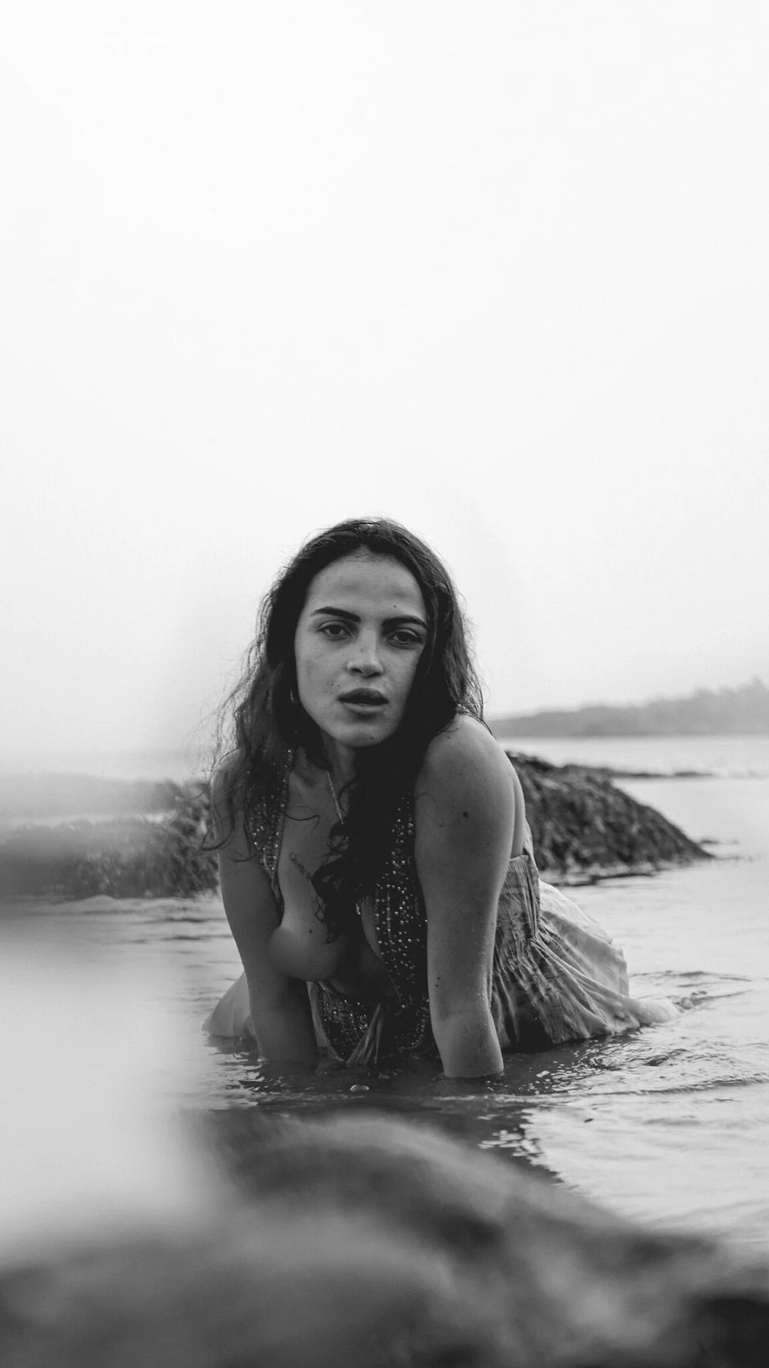 Black and white photo of a woman lying in shallow water on a beach, with rocks and shoreline in the background.