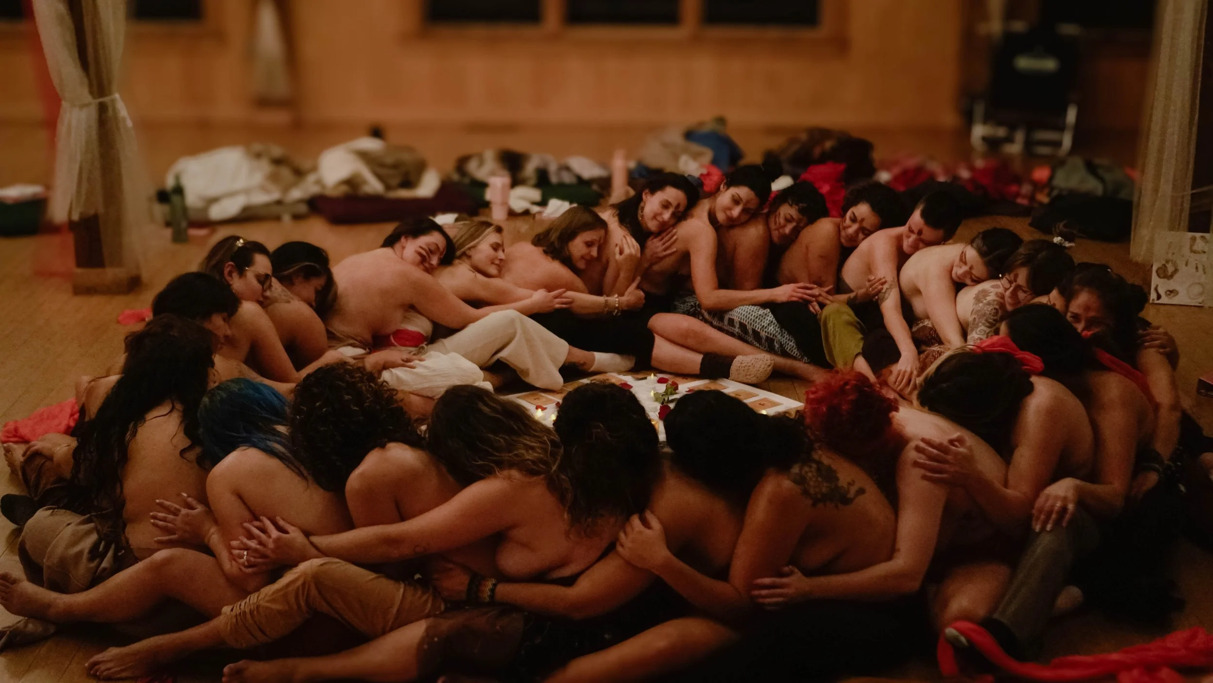 Group of women and girls of diverse ethnicities sitting on the floor in a circle, leaning on each other, smiling, and engaging in a close, affectionate moment inside a wooden room.