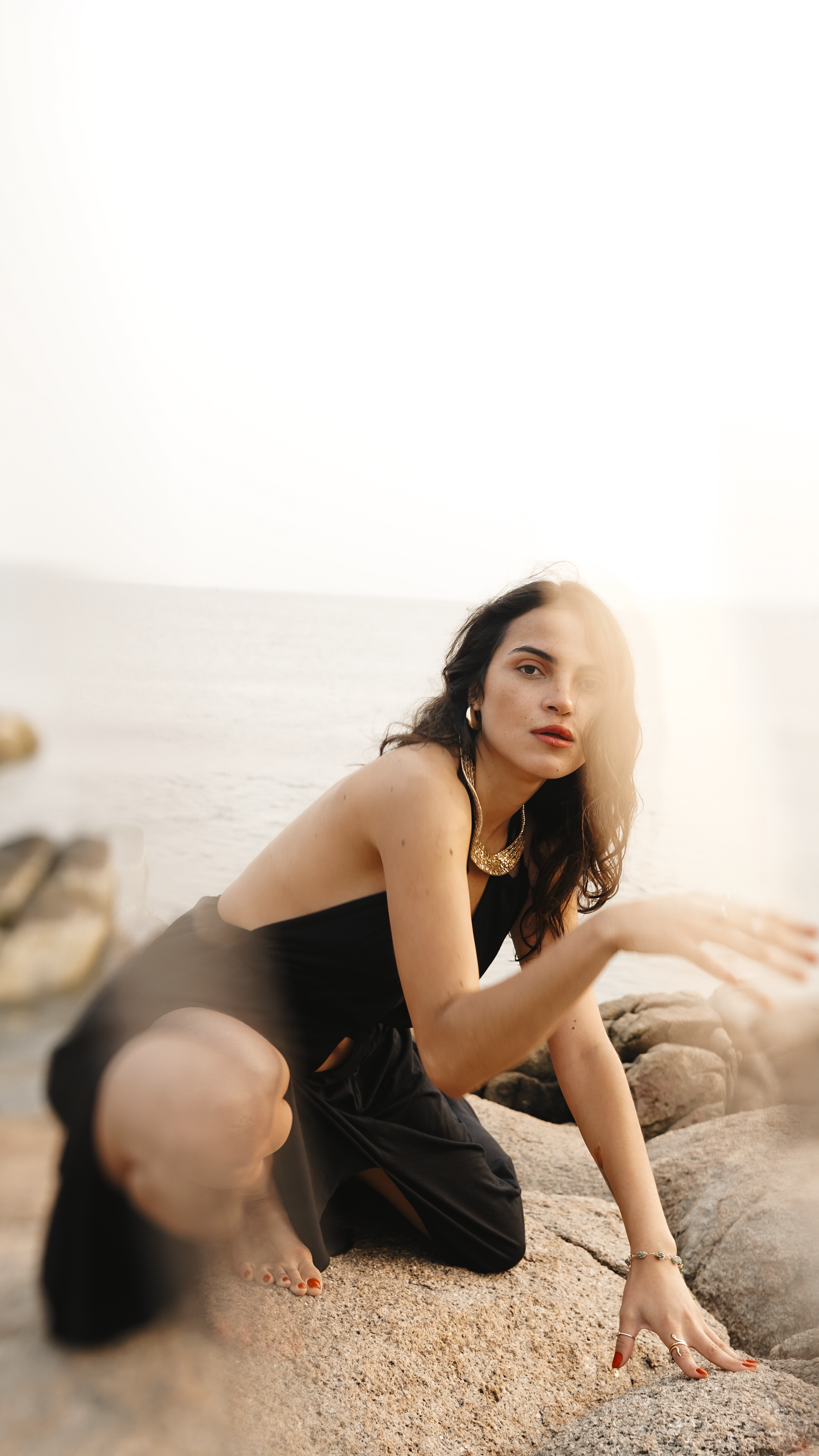 A woman with curly dark hair and light skin crouching on a sandy beach near rocks, wearing a black dress, gold jewelry, and red nail polish, with the ocean and sky in the background at sunset.