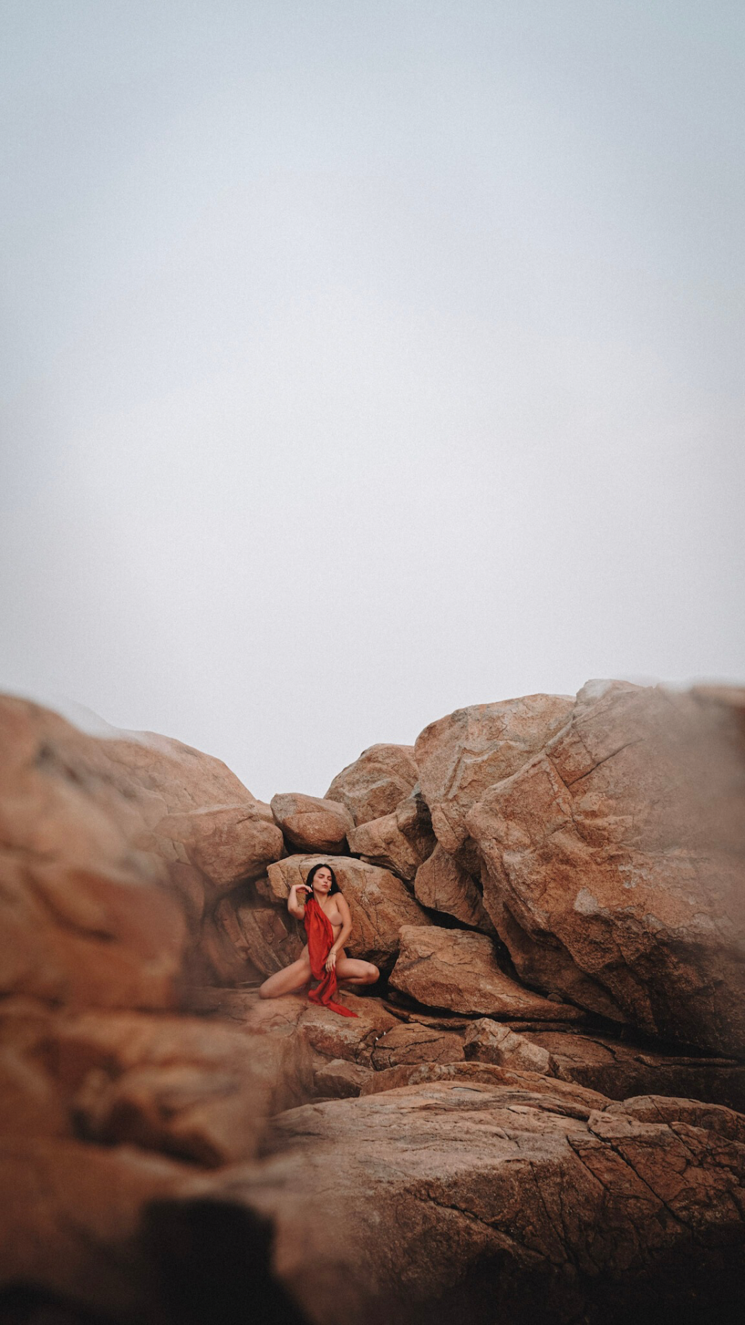 A woman in a red garment is sitting among large rocks in a desert-like landscape, with an overcast sky overhead.