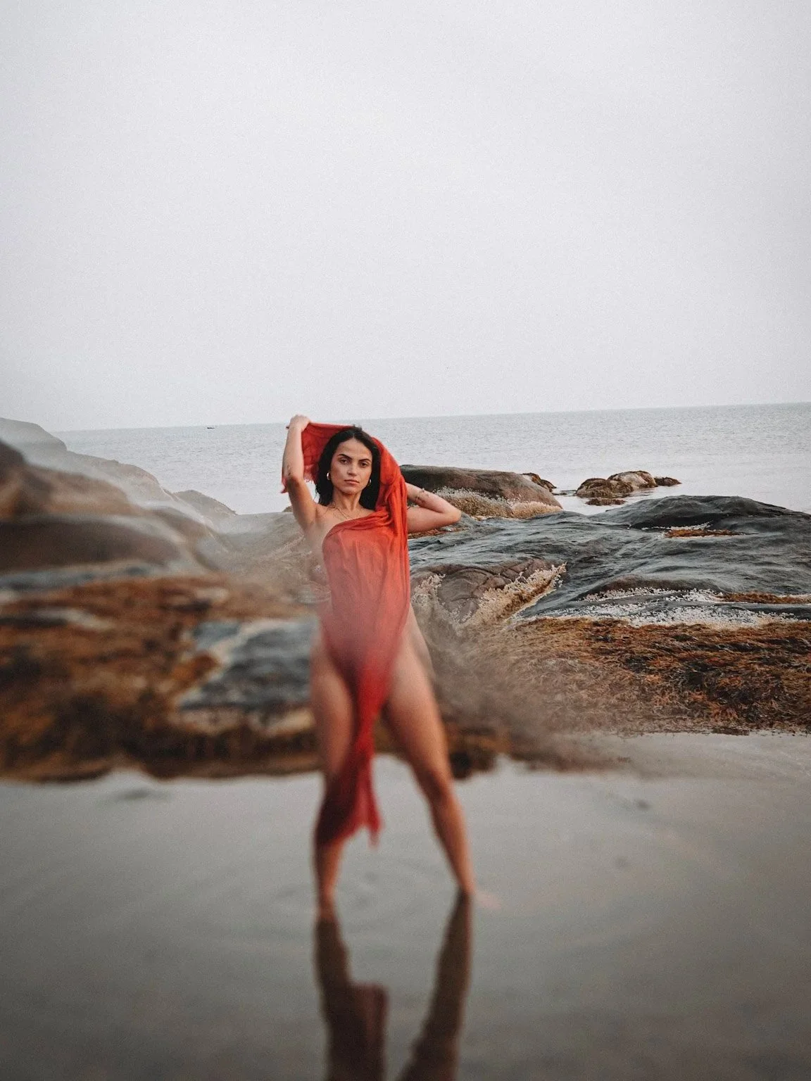 A woman standing on rocks at a beach during sunset or sunrise, holding a red cloth, with the ocean in the background and a cloudy sky.