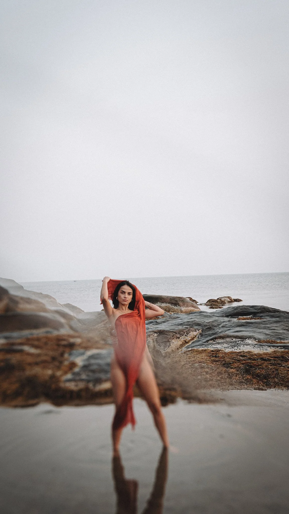 A woman in a red dress standing on rocks by the ocean, with her hand behind her head and the other hand on her hip.