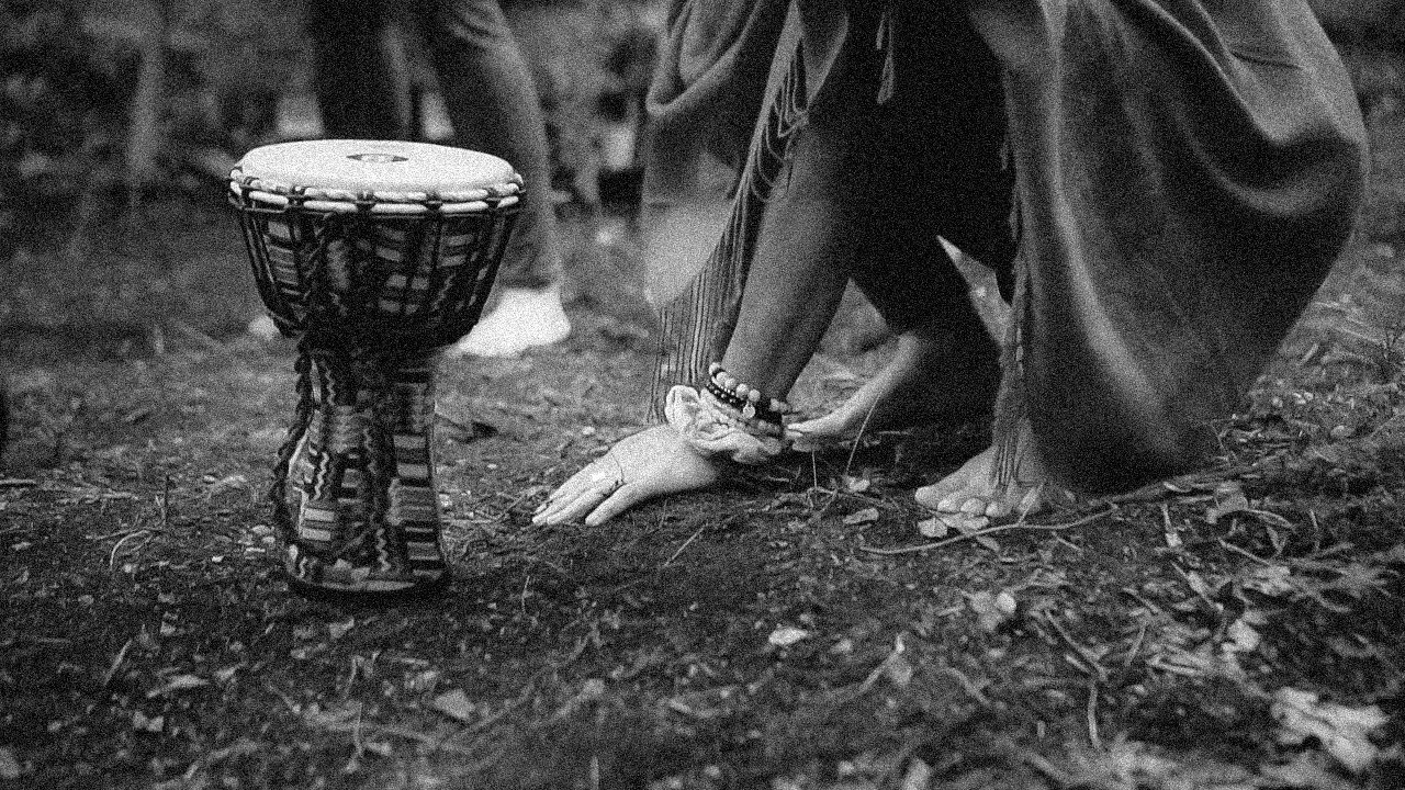 A person is kneeling on the ground with hands on dirt, wearing a long garment and bracelets, near a small drum with a patterned cloth covering, set outdoors on a natural surface.