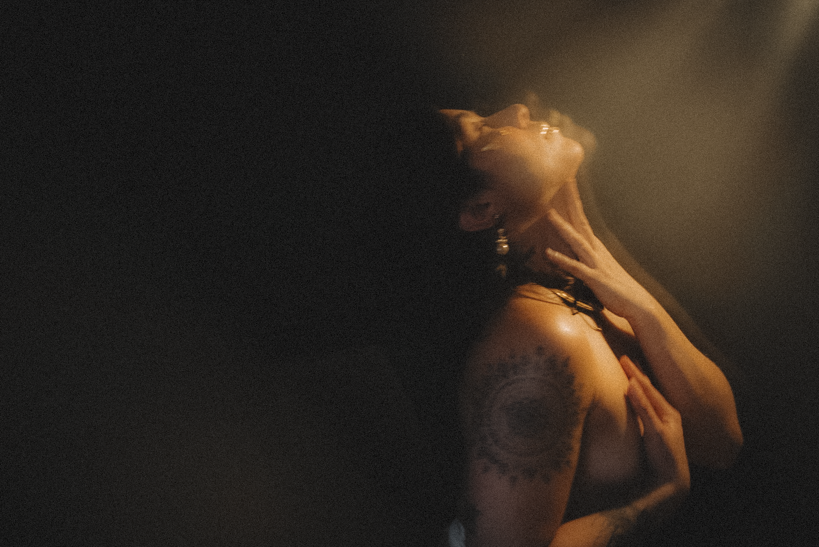 A woman with long dark hair and tattoos on her arm is posing with her head tilted back and her hand touching her neck in a dimly lit setting
