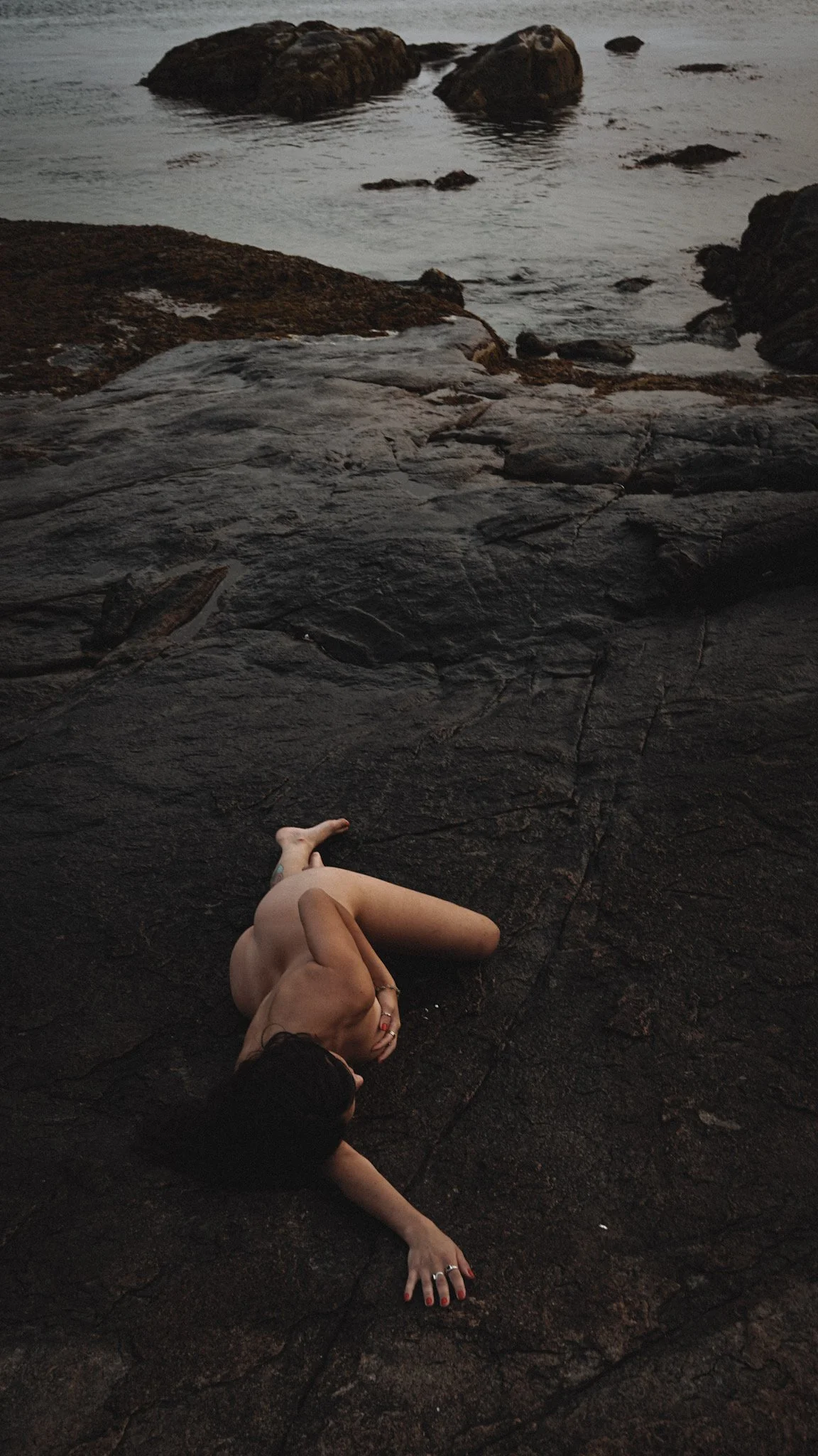 A person lying on a dark, rocky surface near water, with their legs and upper body visible, wearing black nail polish and jewelry, with a view of rocky shoreline and water in the background.