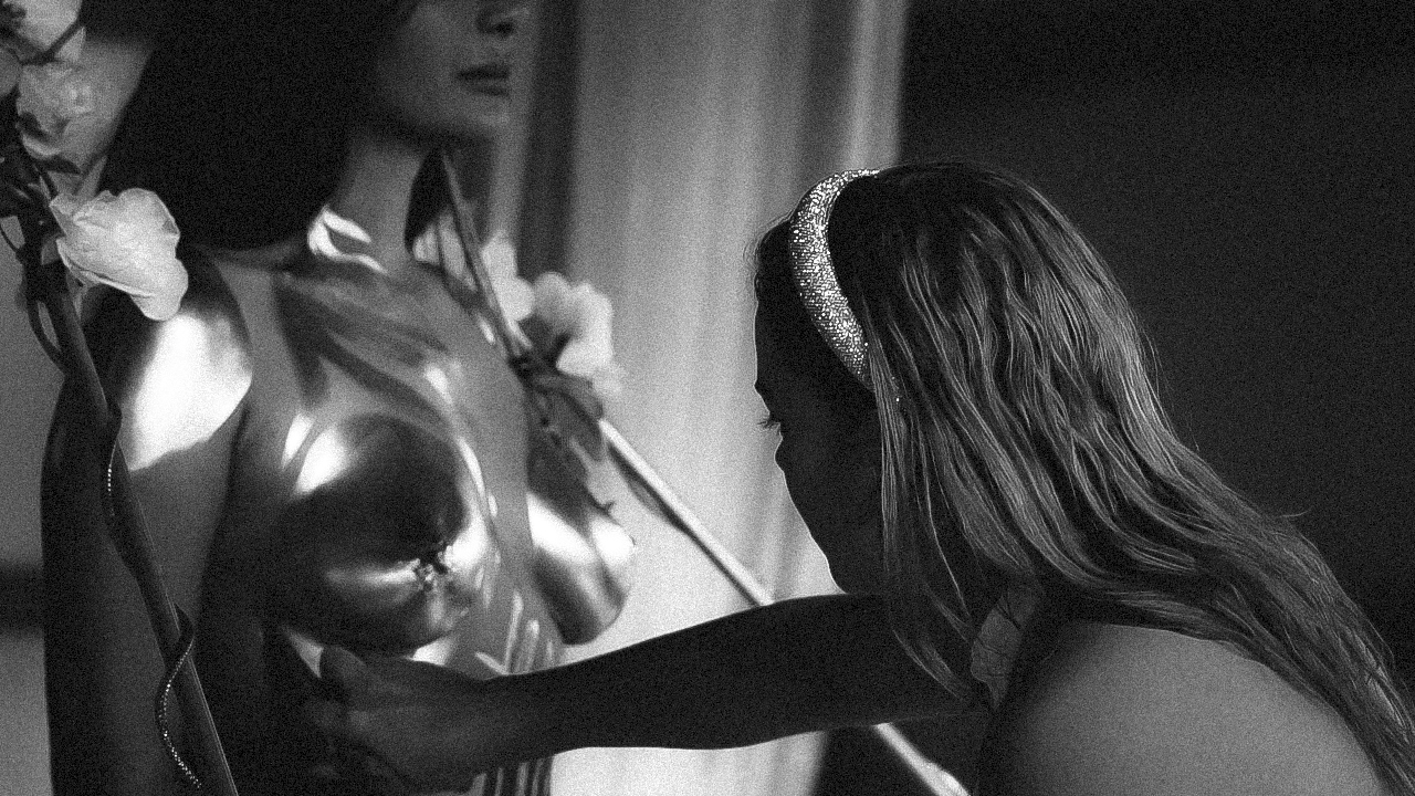 A person with long, wavy hair wearing a shiny headband is kneeling and touching a metallic statue of a woman holding a staff.