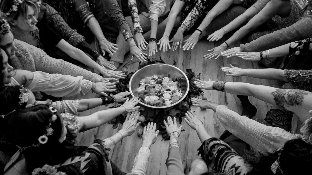 People gathered in a circle with their hands extended toward a bowl of flowers on the floor.