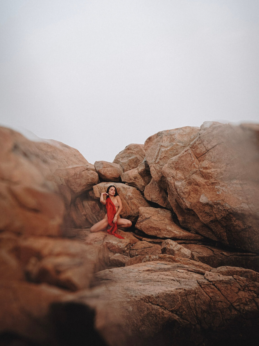 A woman in a red dress sitting among large rocks in a desert landscape under a gray sky.