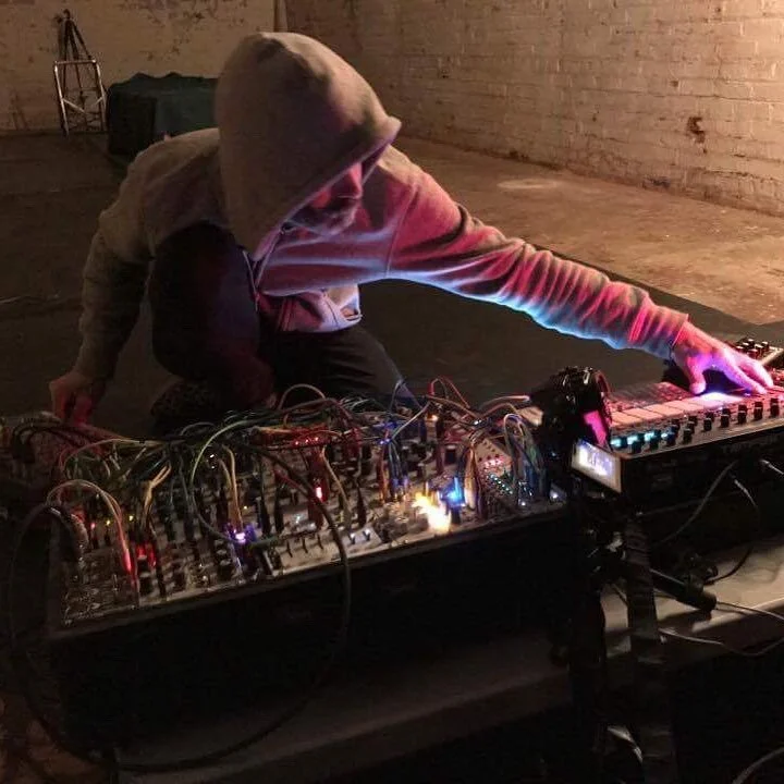 Person wearing a hoodie, kneeling and adjusting electronic music equipment with various cables in a dimly lit, rustic room.