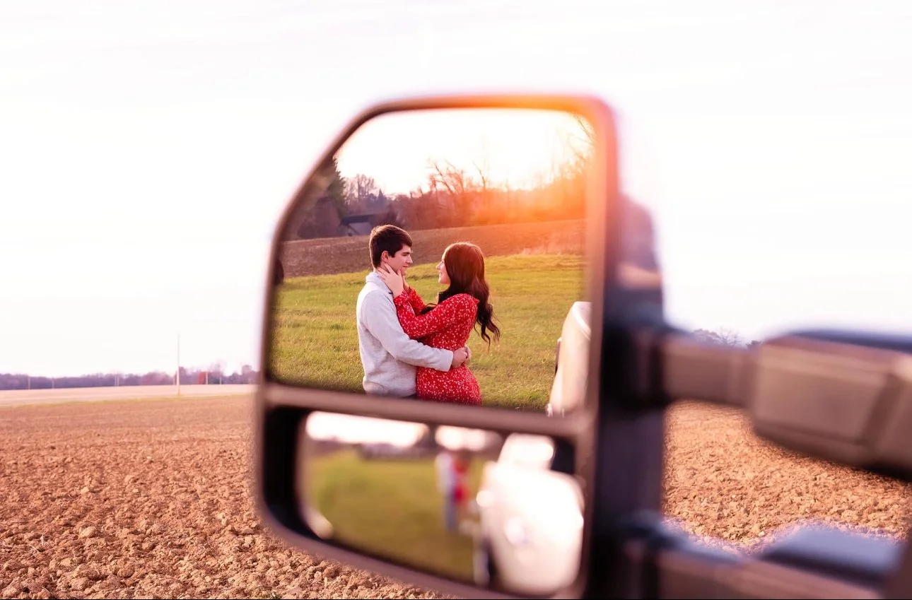 A couple embracing in a field, seen through a truck's side mirror. The sunset casts a warm glow over the scene.