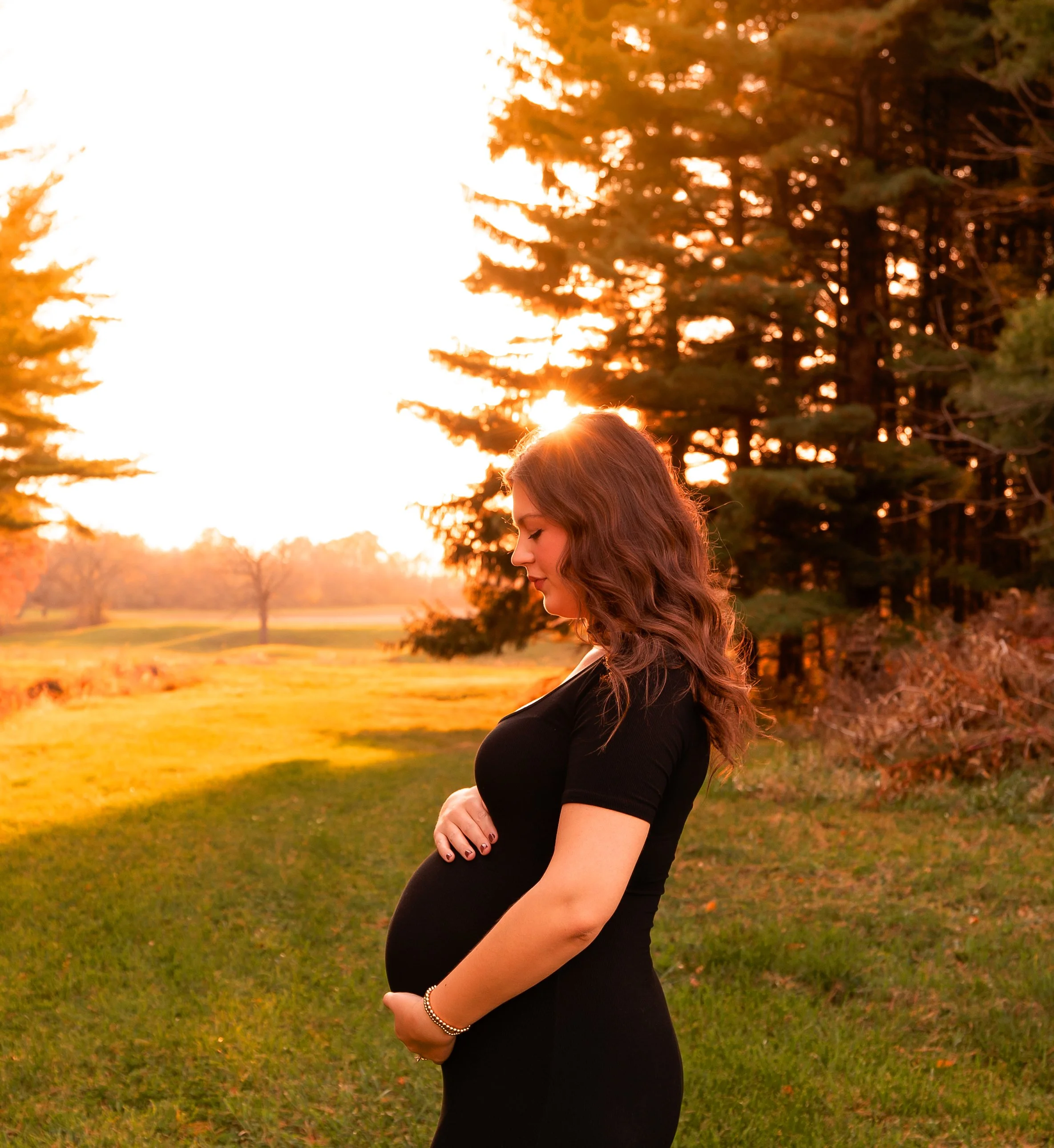 Pregnant woman in black dress standing in a grassy field during sunset, gently cradling her belly with both hands, facing sideways with a peaceful expression.