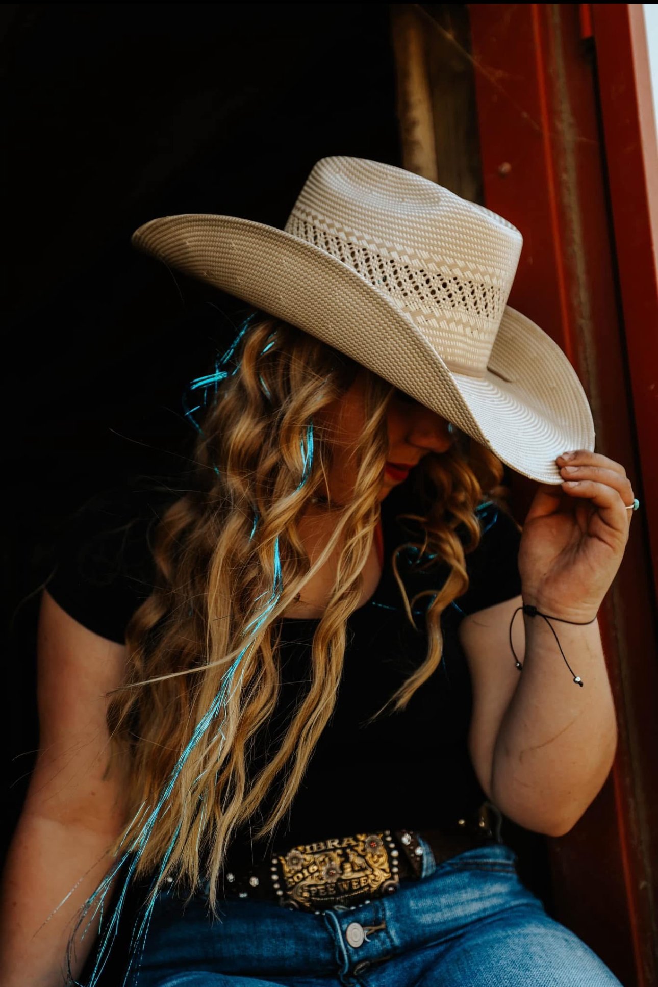 A woman with long wavy hair wearing a large straw hat, a black shirt, a decorative belt, and blue jeans, leaning against a wooden wall.