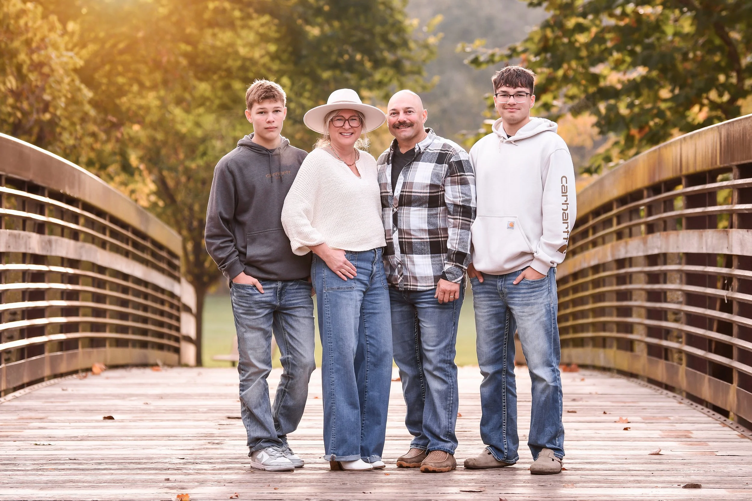 Family of four standing on a wooden bridge outdoors during fall, surrounded by trees with autumn foliage, smiling at the camera.